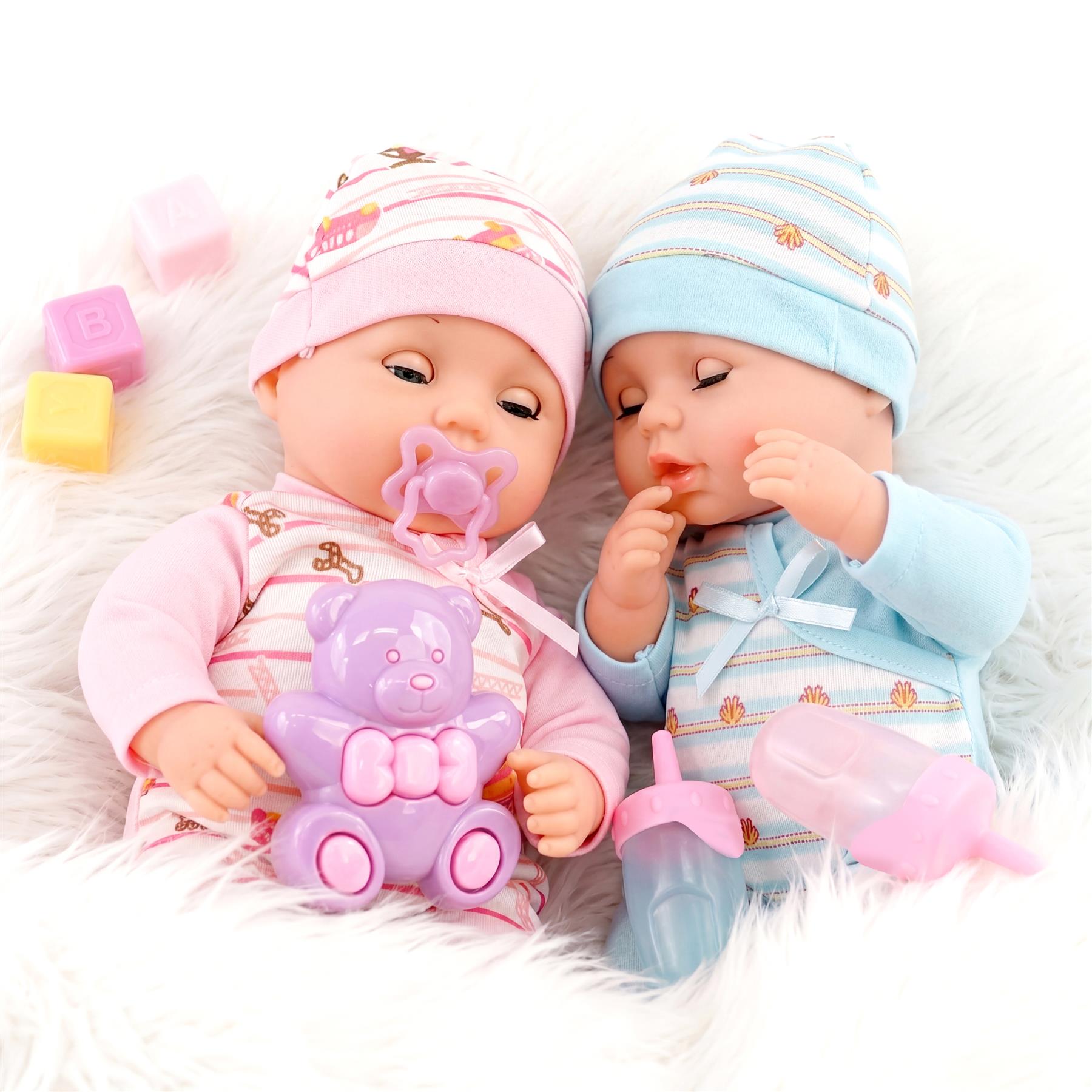 Twin dolls dressed in pink and blue outfits rest with a teddy bear, bottles, and colorful accessories.