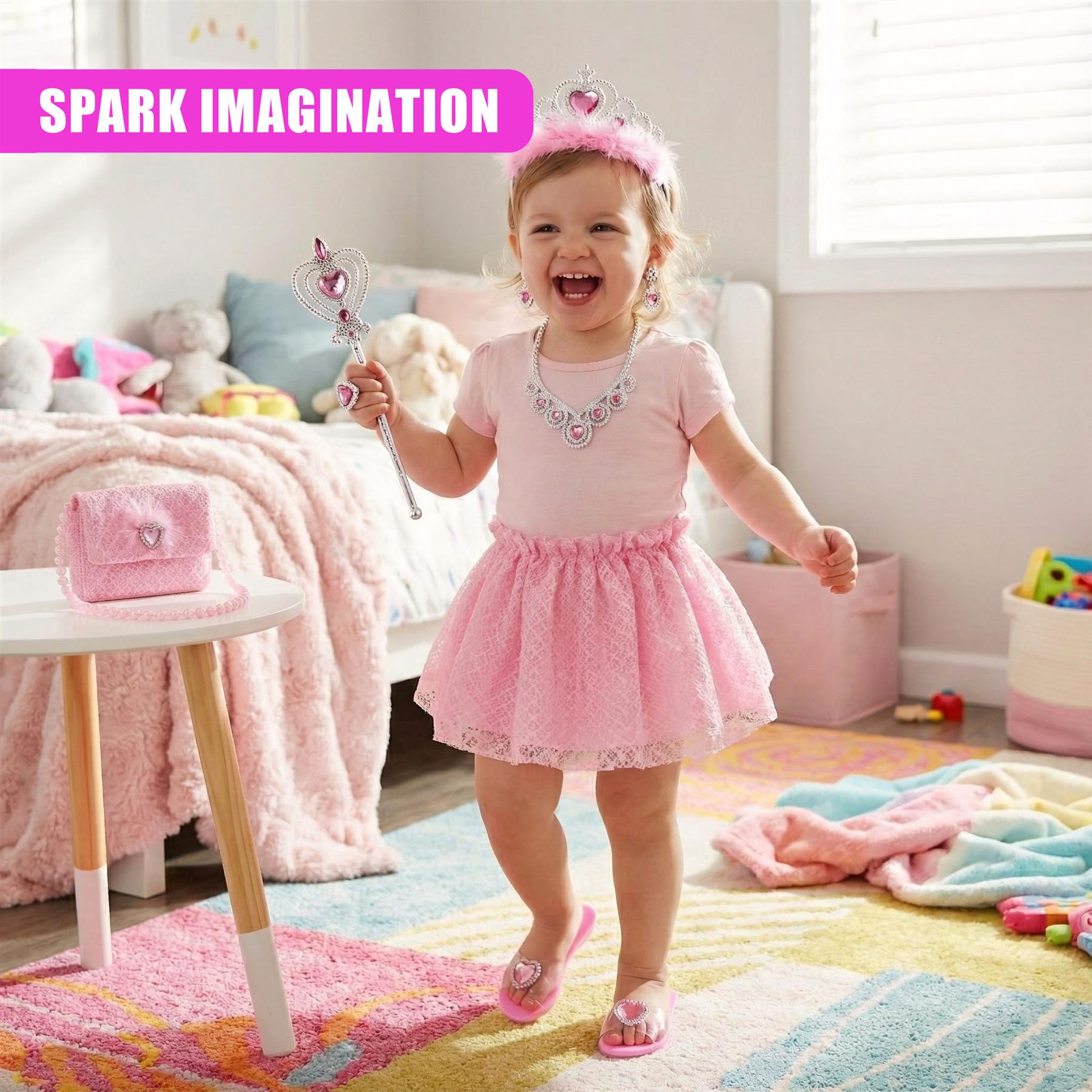 A young girl, around 2-3 years old, is shown in a playroom, wearing a pink tutu and a tiara. She's holding a sparkly wand and has a big smile on her face. The room is filled with toys and a bed in the background, with a few toys and a stuffed animal on the bed. The child is standing on a colorful rug, surrounded by toys and a play area. - The Magic Toy Shop