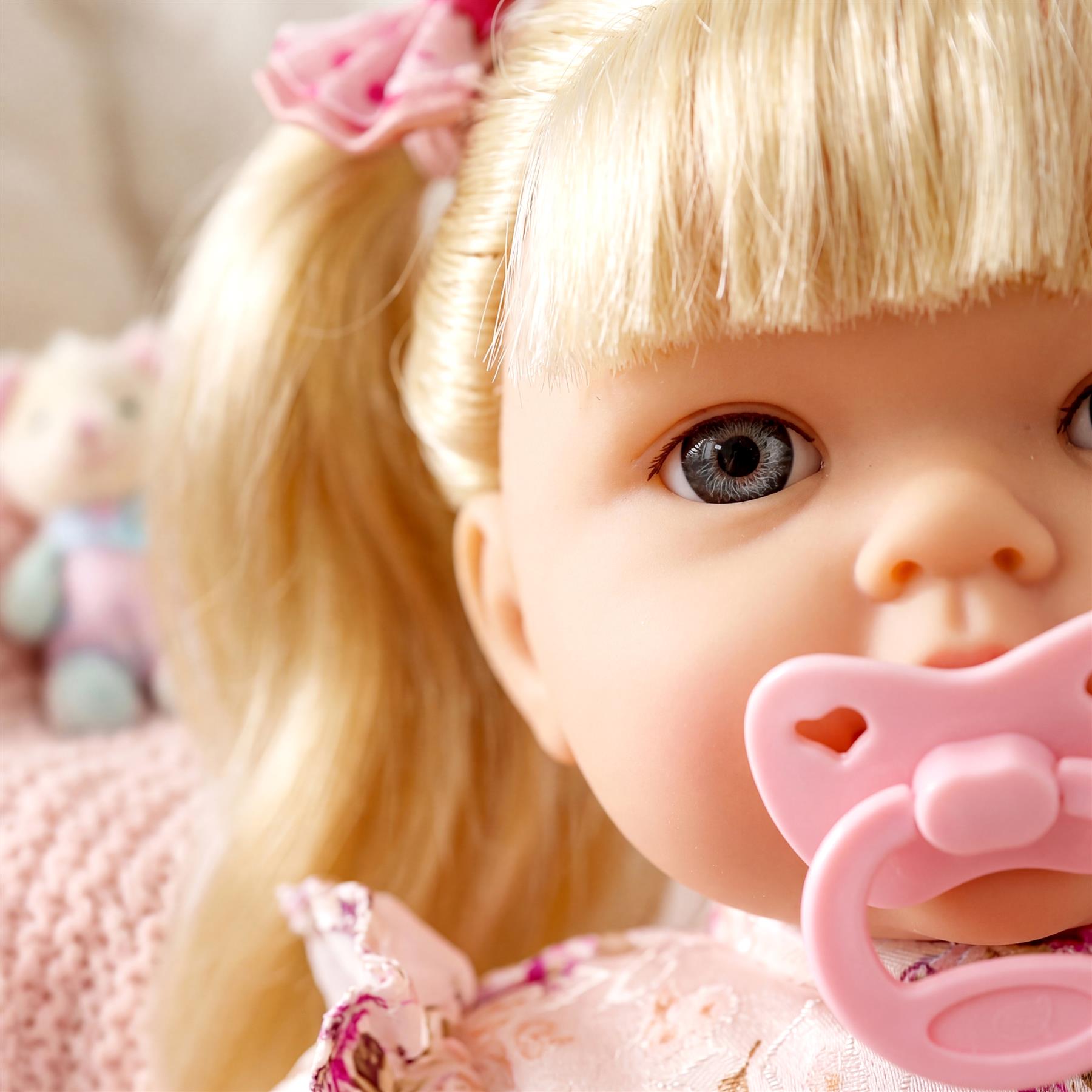 Blonde doll with blue eyes and light pink outfit, holding a pale pink pacifier.