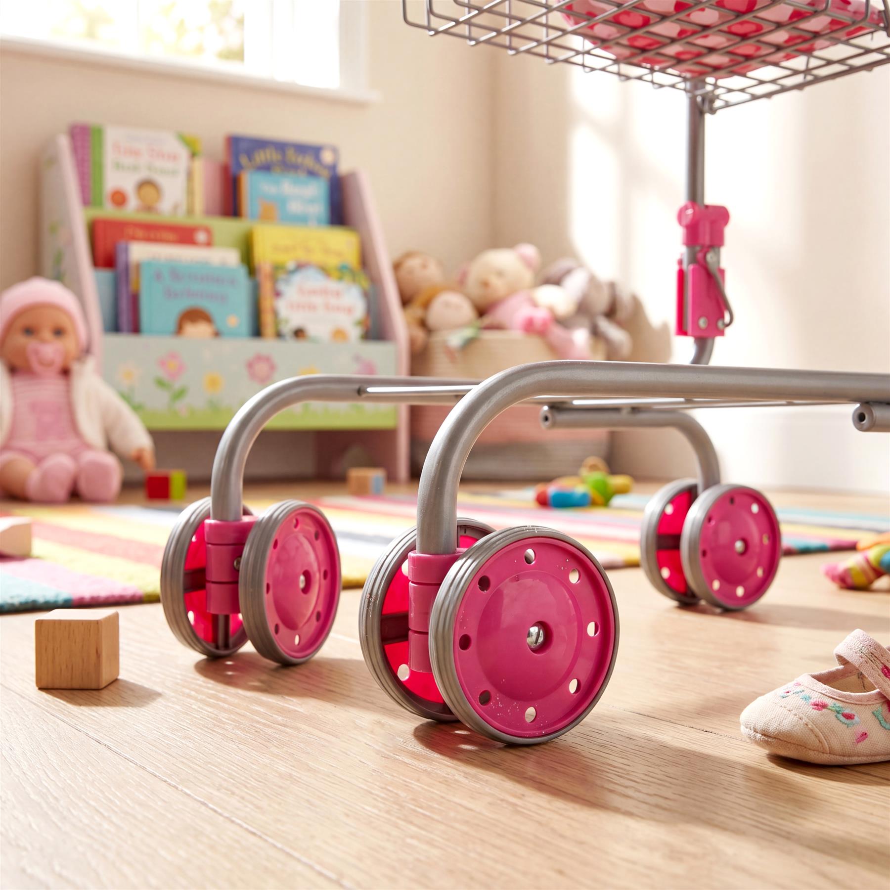 A miniature silver shopping cart with four pink wheels sits on a wooden surface near books.