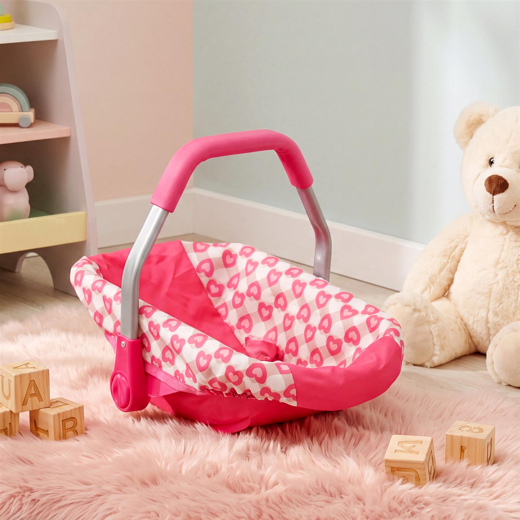 Pink baby car seat with handle rests on a fuzzy pink surface near a bear stuffed animal.