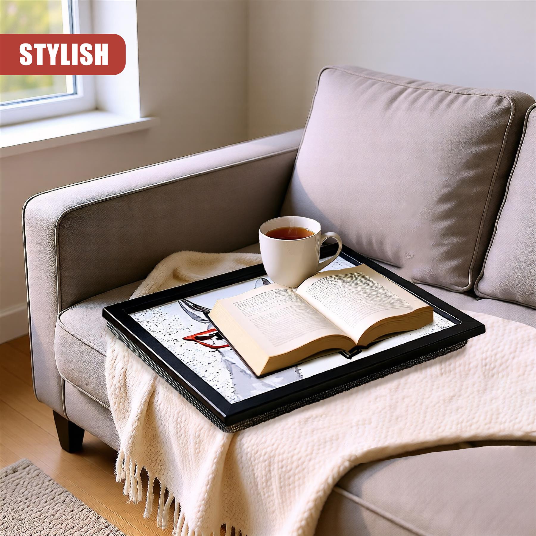 A black tray holding an open book and white mug rests on a beige throw blanket on a sofa.
