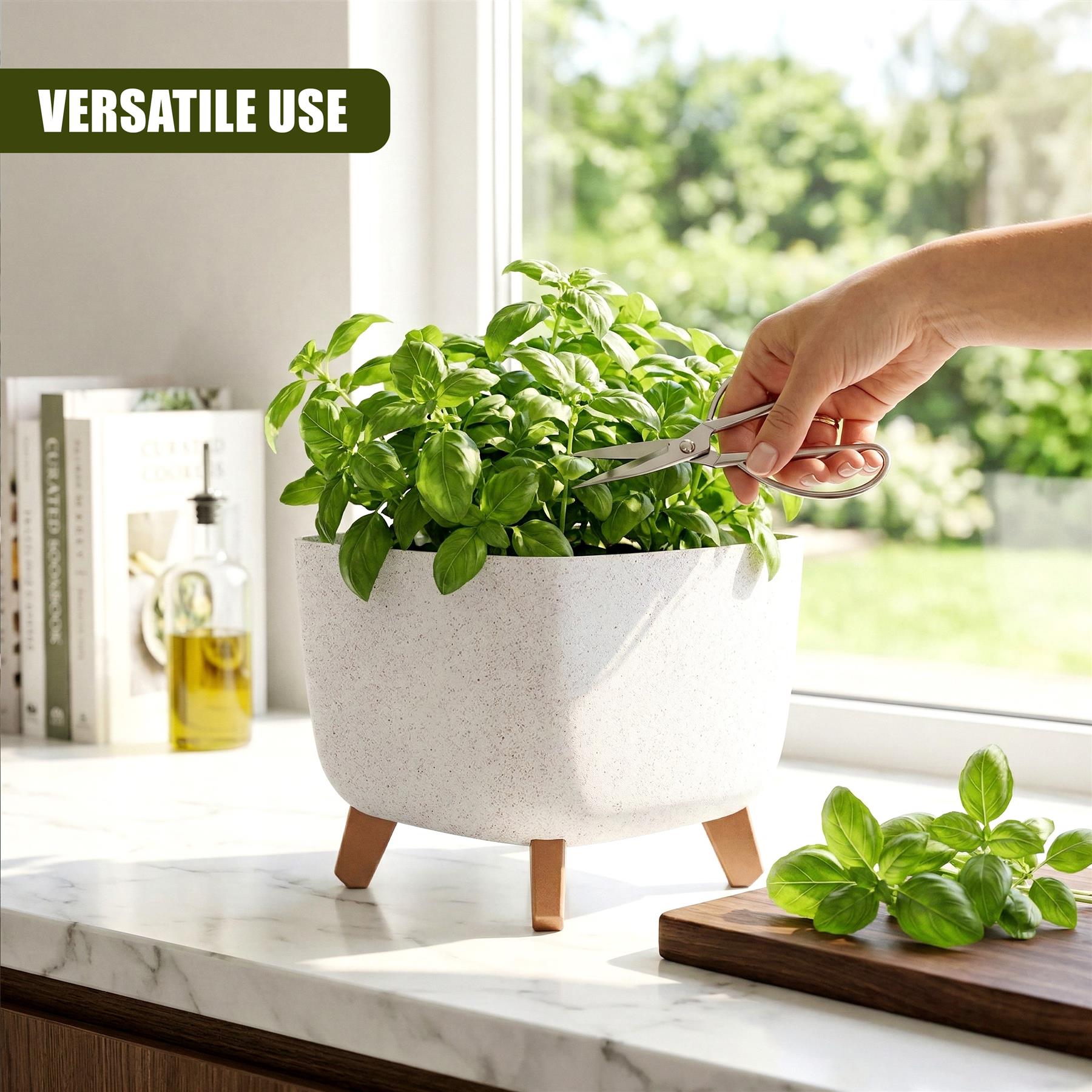 Basil plants grow in a speckled white pot with legs; a hand uses scissors to trim leaves.