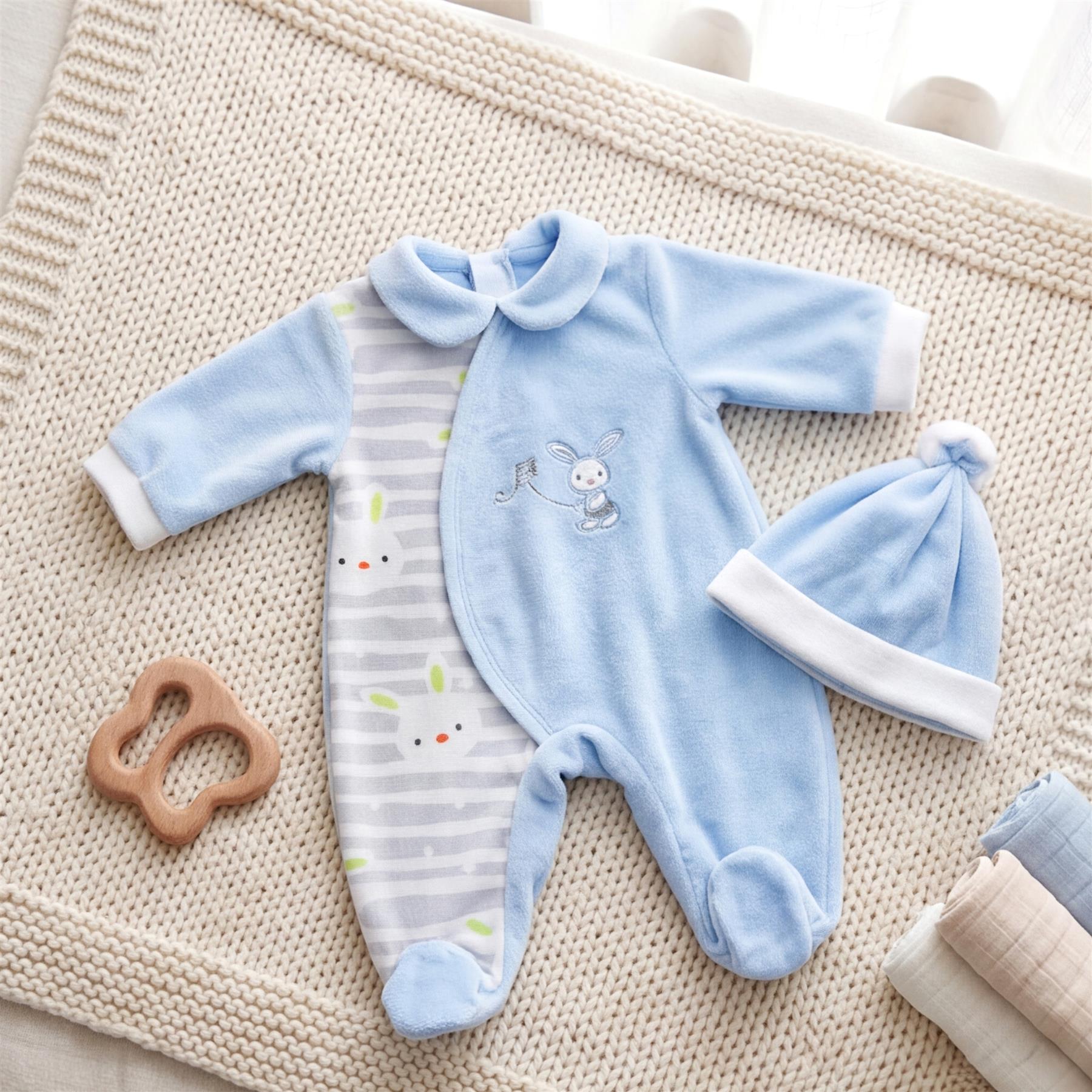 A light blue doll clothes set includes a romper, hat, and striped garment, next to a wooden ring.