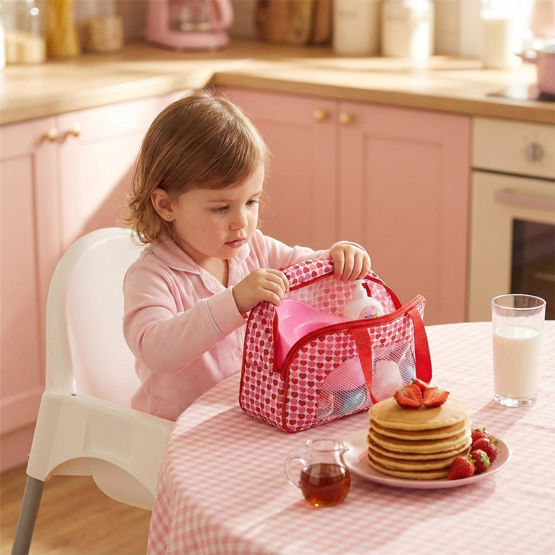 A young girl sits at a table in a kitchen, surrounded by a breakfast spread. She holds a pink and white lunch bag with a handle, which is open to reveal a pink cup, a white cup, and a blue cup. A plate of pancakes with strawberries is in front of her, and a glass of milk is to the right. The tablecloth is a red and white checkered pattern. - The Magic Toy Shop