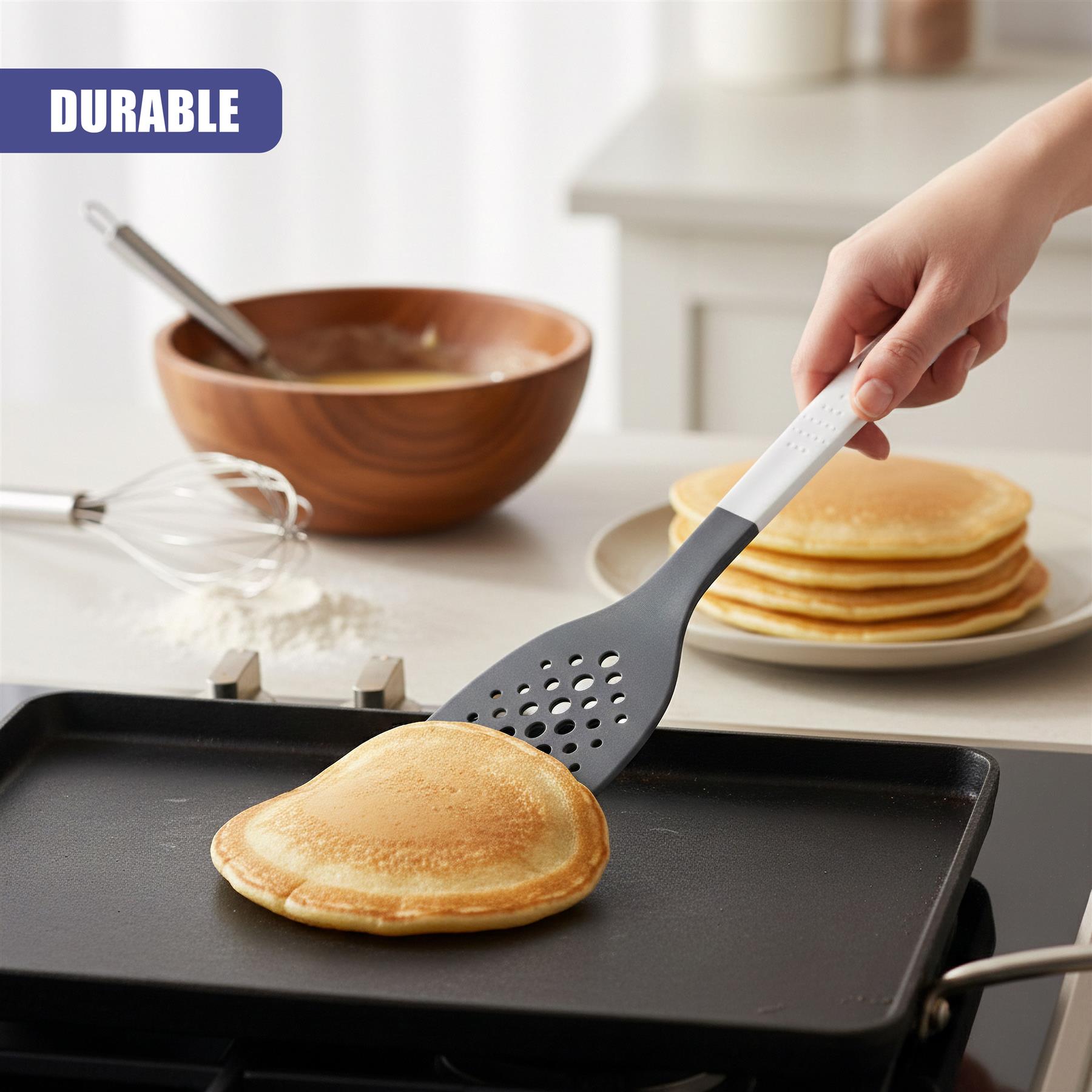 Perforated spatula lifts a pancake over a black griddle near stacked pancakes and flour.