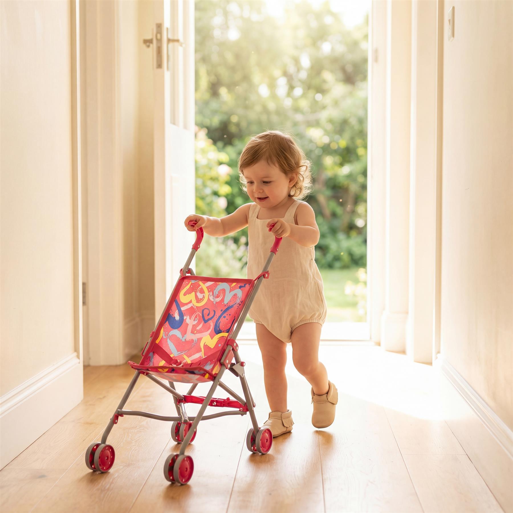 Toddler holding a patterned pink toy stroller indoors, standing on light wooden floors.