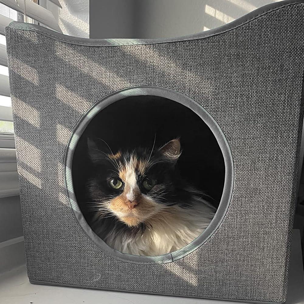 A grey, fabric cat house with a large, round window. A calico cat with white, black, and orange fur is visible through the window, looking directly at the camera. - The Magic Toy Shop
