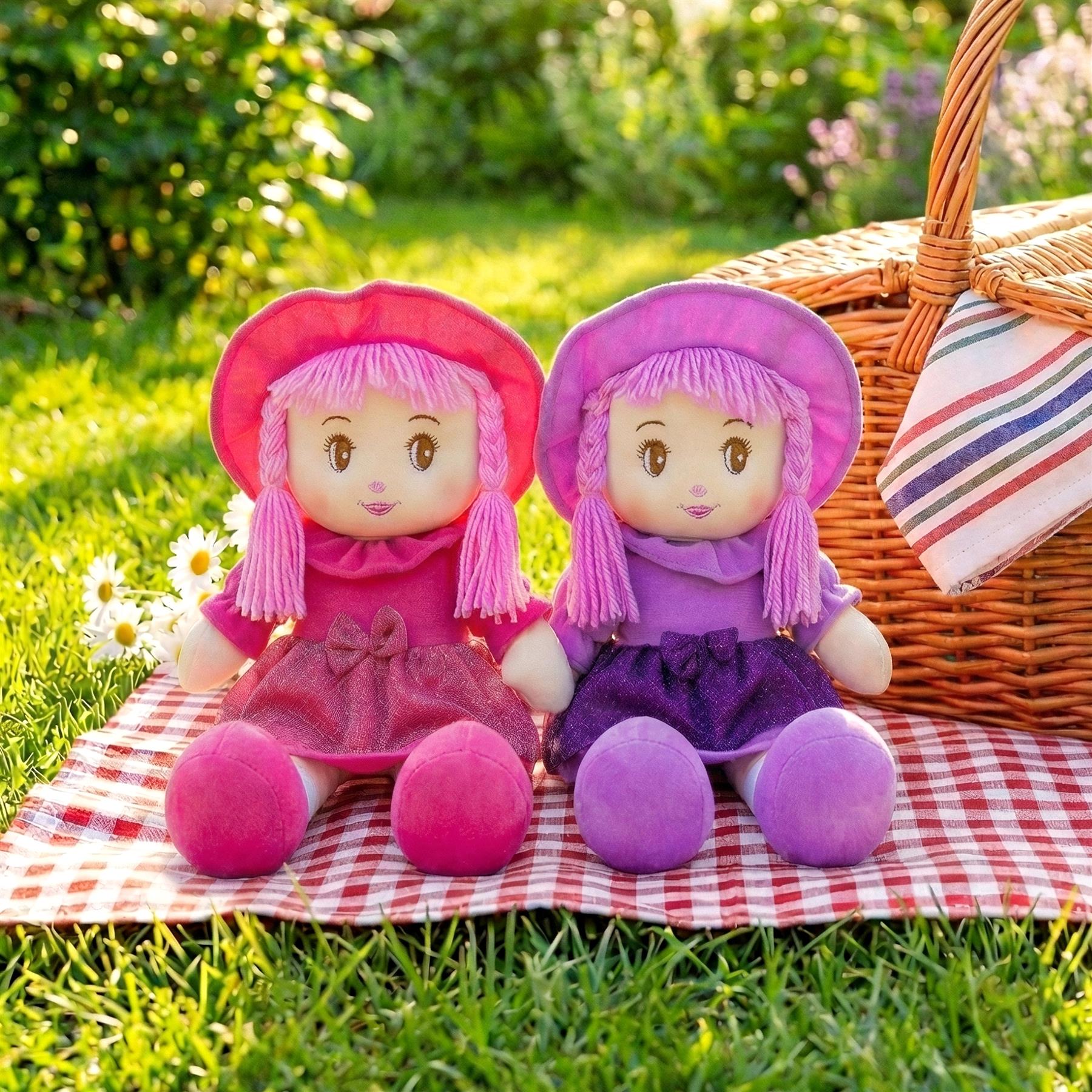 Two rag dolls, one pink and one purple, sit on a red checkered blanket near a woven basket.