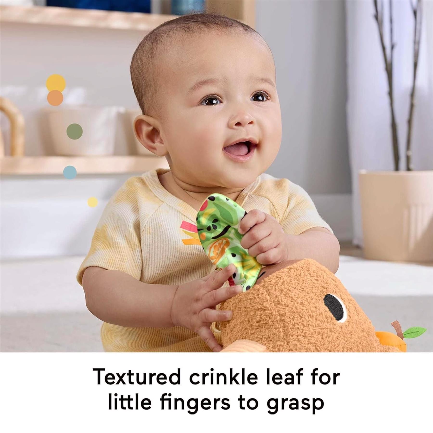A baby holding a textured, crinkle leaf attached to an orange capybara plush toy.