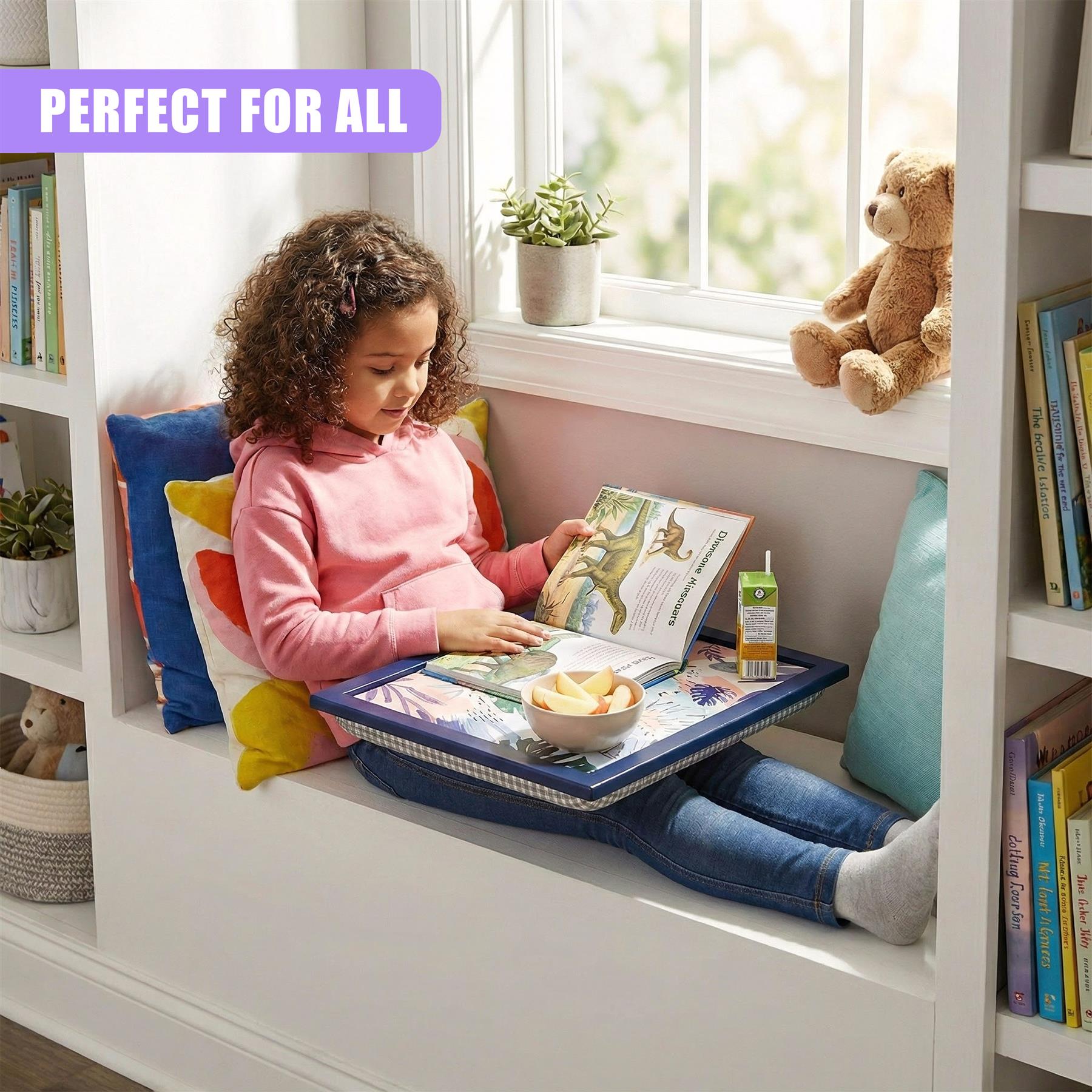 Young girl using a blue lap tray with a leaf pattern while reading an open book on a window seat.
