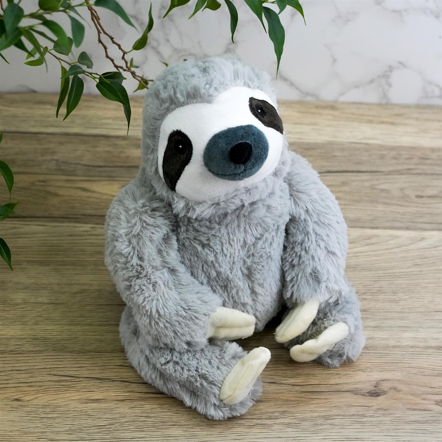 A soft, grey plush toy shaped like a sloth, sitting on a wooden surface. The sloth has a white face with black eyes and a blue nose. It has long, grey arms and a big, grey body. It is facing the camera with its arms held out to the sides. The background is a light wood floor and a white wall with a few leaves from a plant in the corner. - The Magic Toy Shop