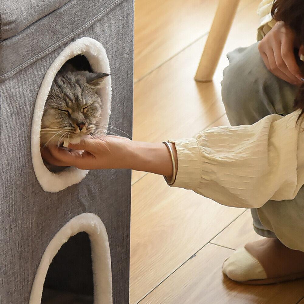 A cat is in a small, gray, two-story cat house. The cat is in the top hole, with its head and paws sticking out. The cat is a tabby, with a gray and brown coat. The cat's eyes are closed, and it is purring. The cat house has a small, white, fluffy rim around the holes. The cat house is made of a gray, fabric-like material, and has a small, gray, fabric-like roof. - The Magic Toy Shop