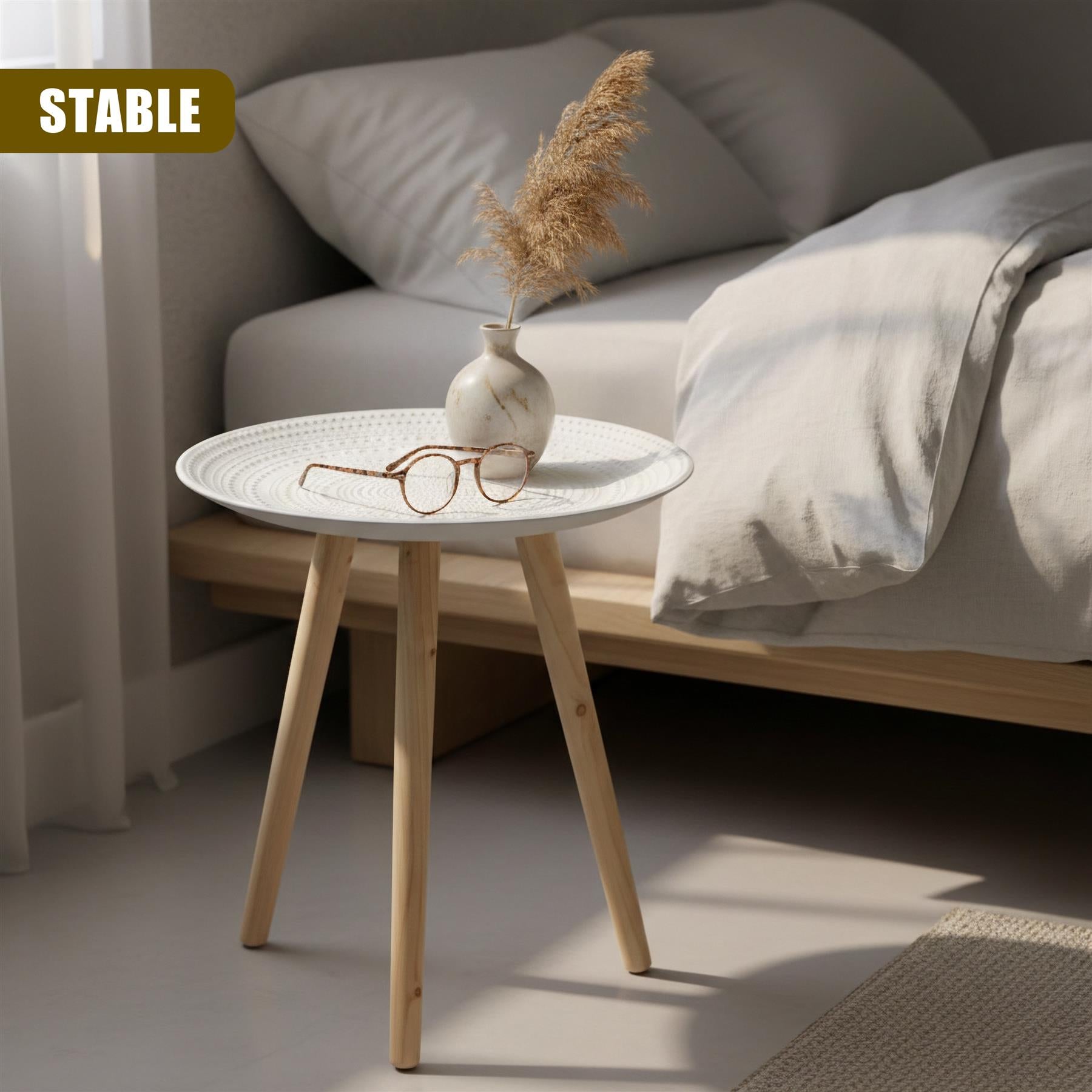 Round white side table with wooden legs holds a vase of dried grass and a pair of eyeglasses.