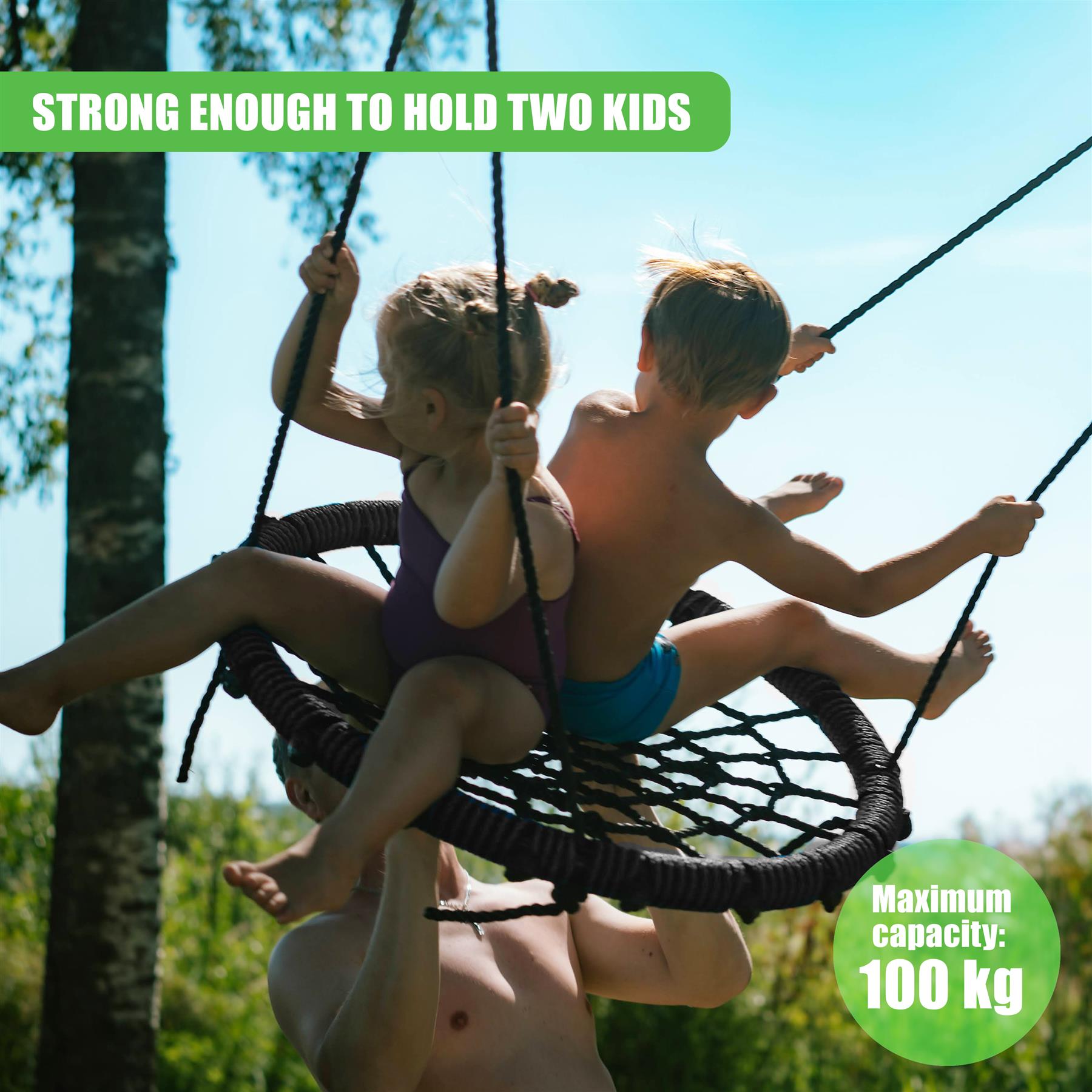 Two children sit in a woven, dark hanging swing suspended outdoors near green trees.