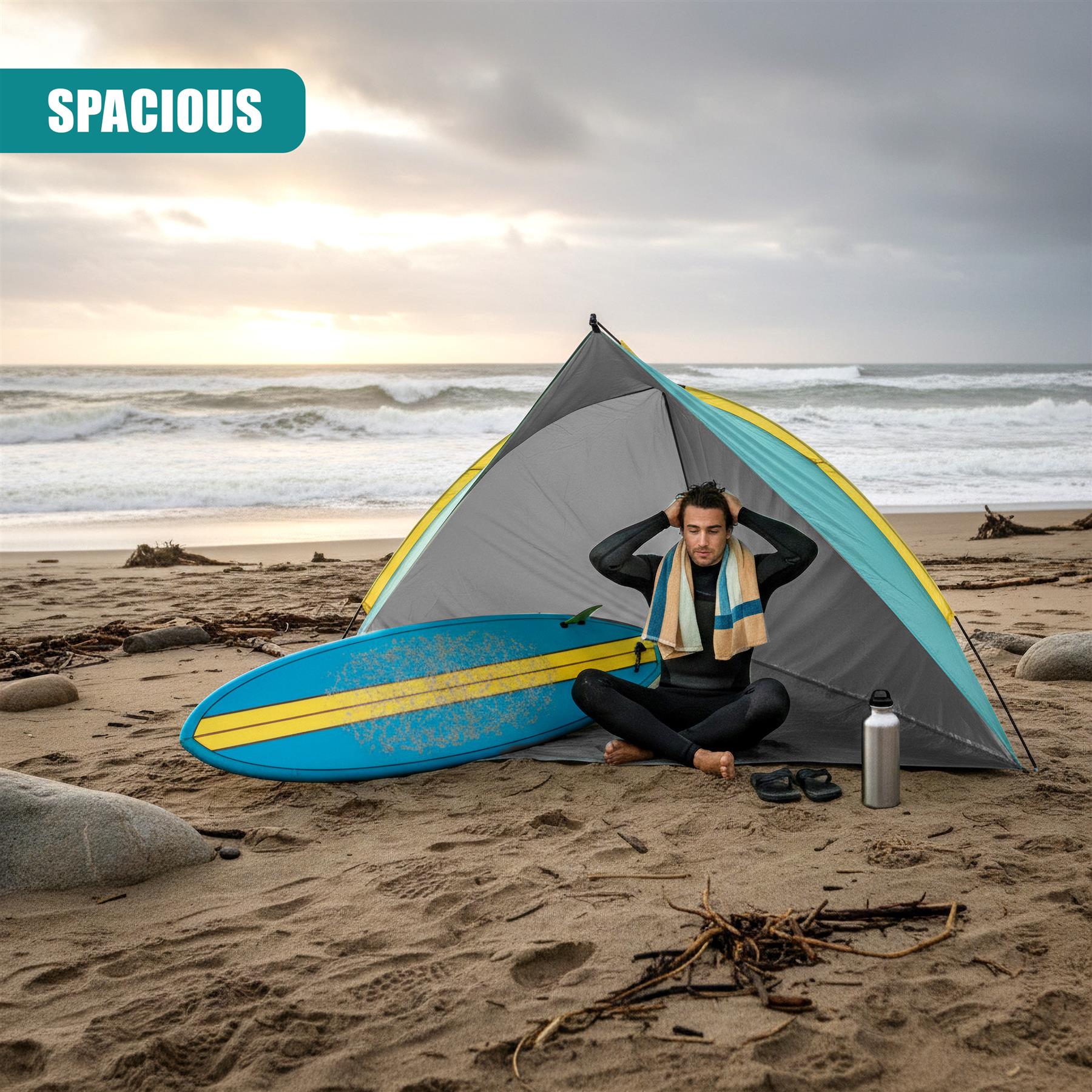 A man sits outside a pop-up tent on the beach with a blue surfboard nearby.