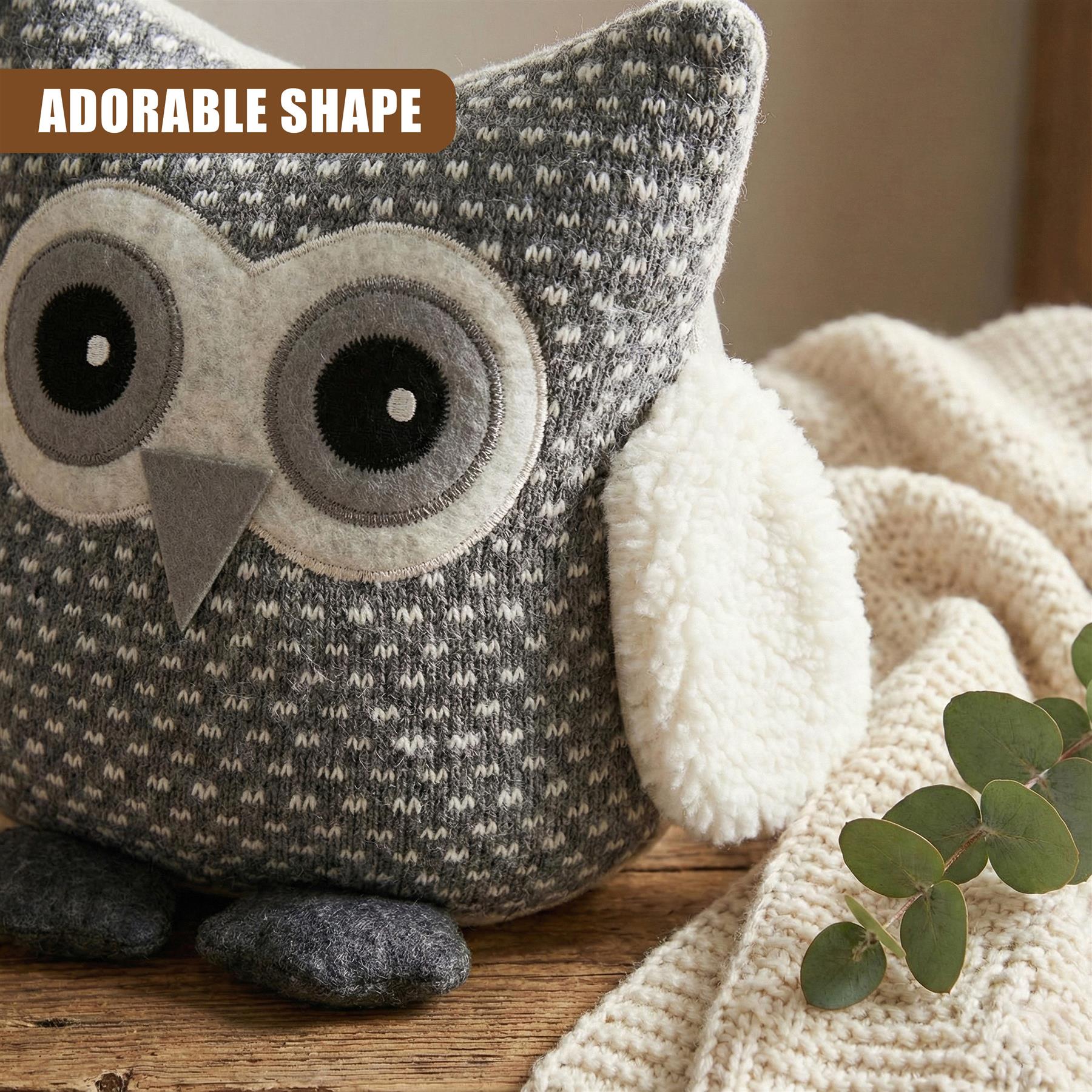 A grey and white owl plush toy with a big, round face and large, round eyes. It has a soft, fluffy body and a small, flat tail. The background is a wooden table with a soft, cream-coloured blanket and a few leaves. The owl is sitting on the table, with its body slightly turned to the right. - The Magic Toy Shop