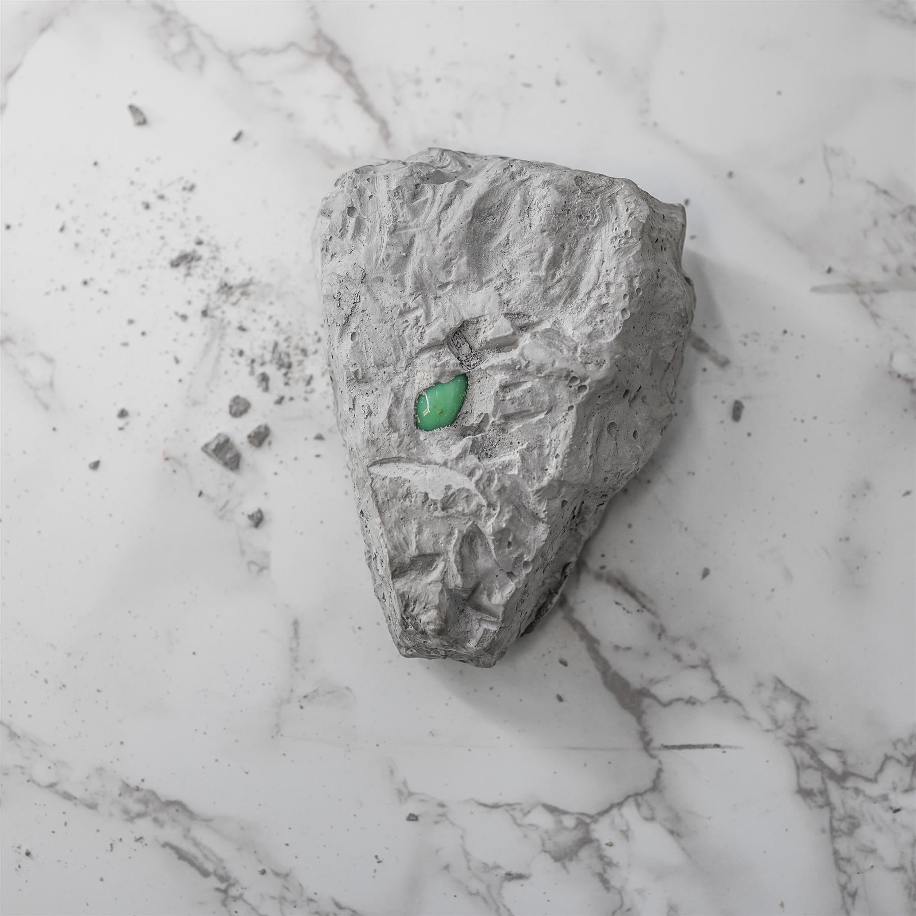 Rough gray rock displayed on white marble surface, featuring a small, smooth green gem.