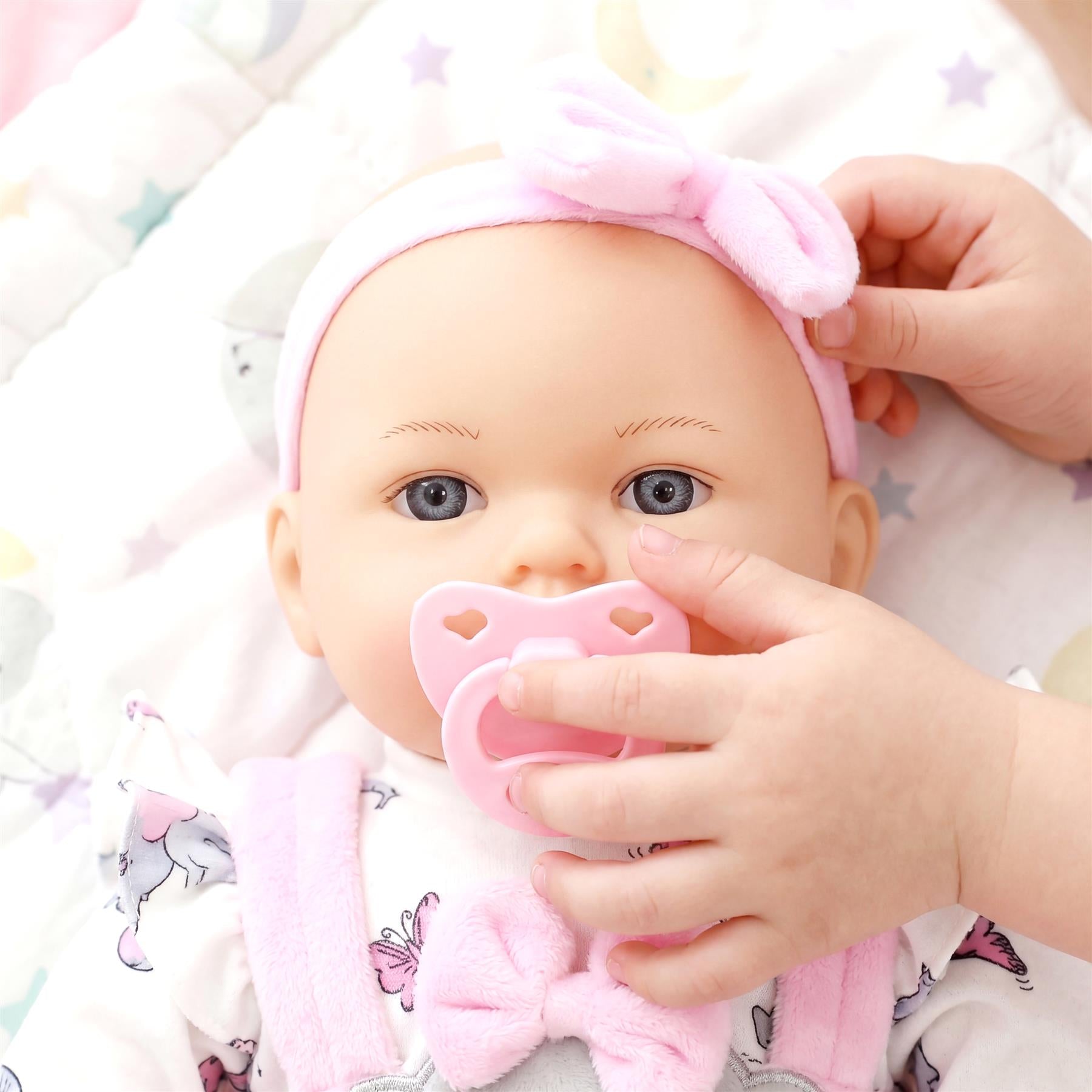 Hands gently place a pink pacifier into the mouth of a baby doll wearing a matching pink headband.