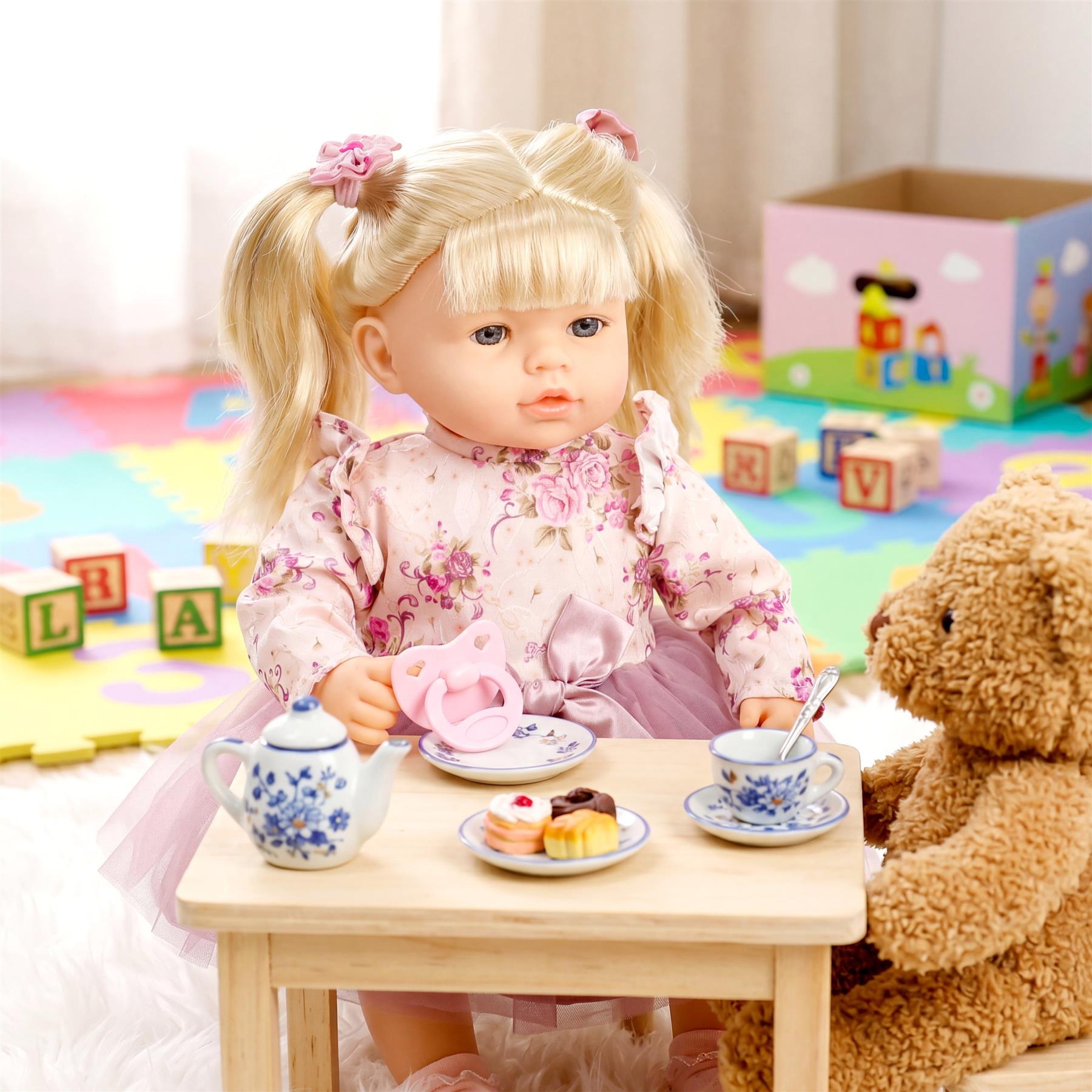 Blonde doll sits at a table with miniature teaware and treats, accompanied by a teddy bear.