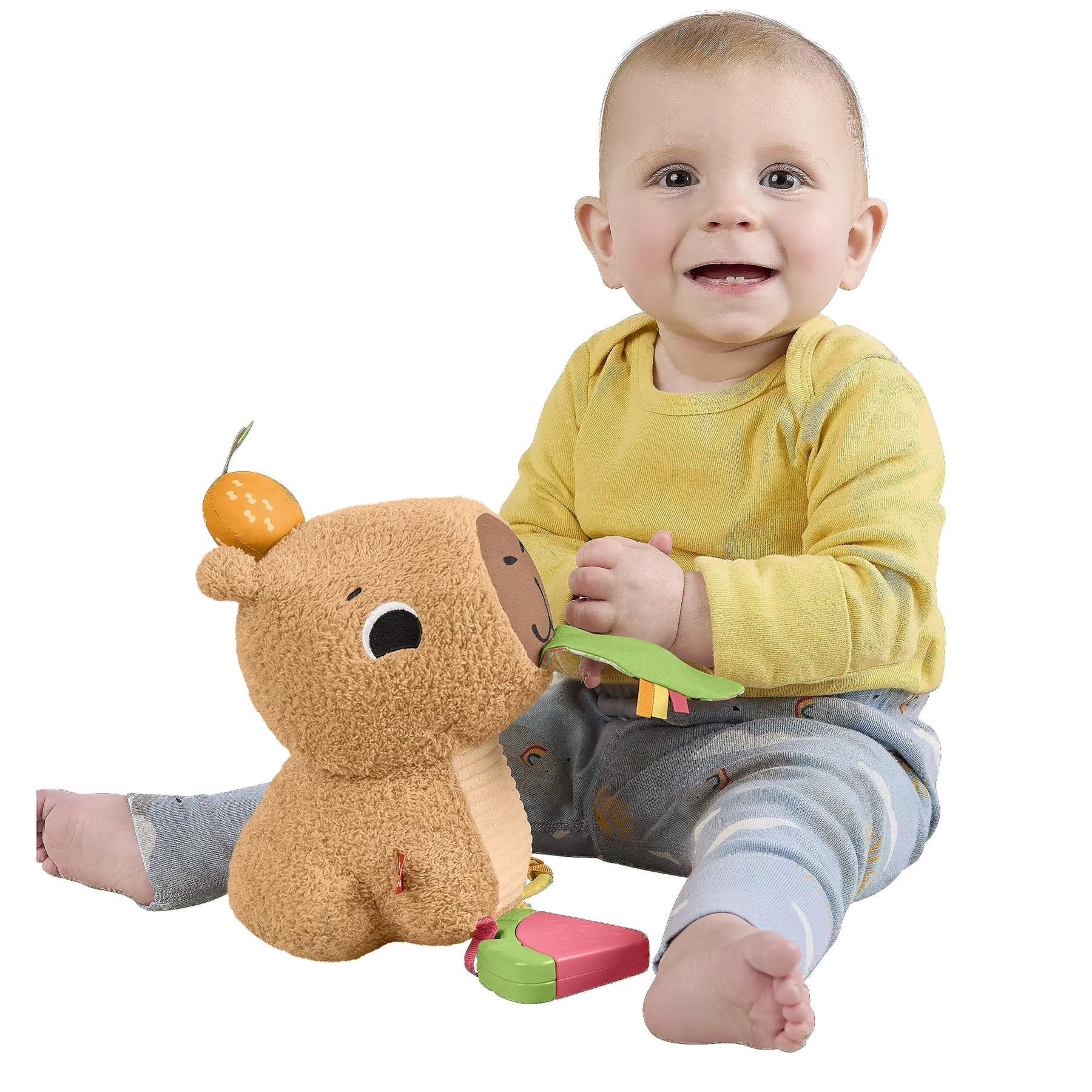 A baby wearing a yellow shirt sits with a brown stuffed capybara plush toy and an orange snack.