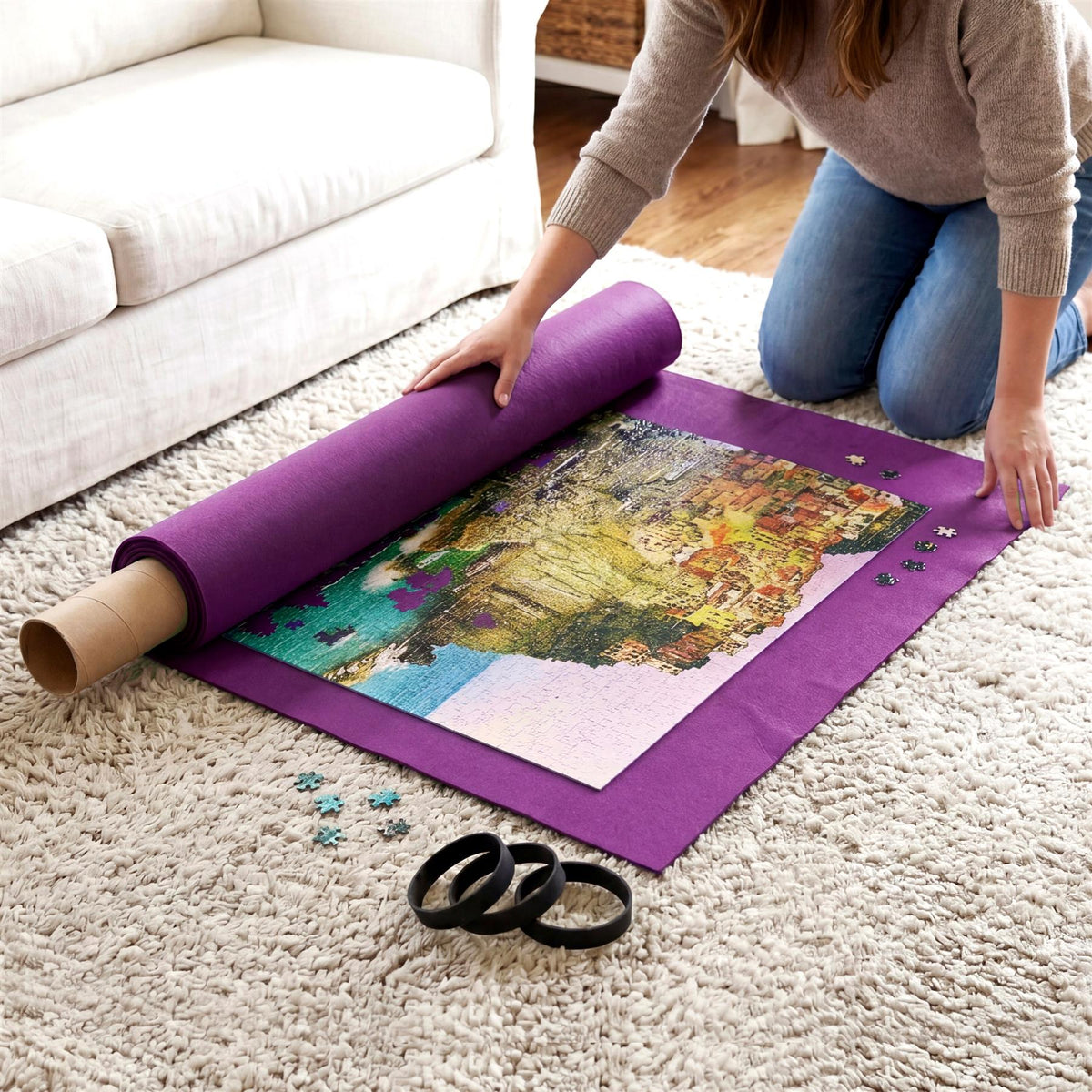A woman kneels on a rug, rolling up a large purple jigsaw puzzle mat with scattered pieces