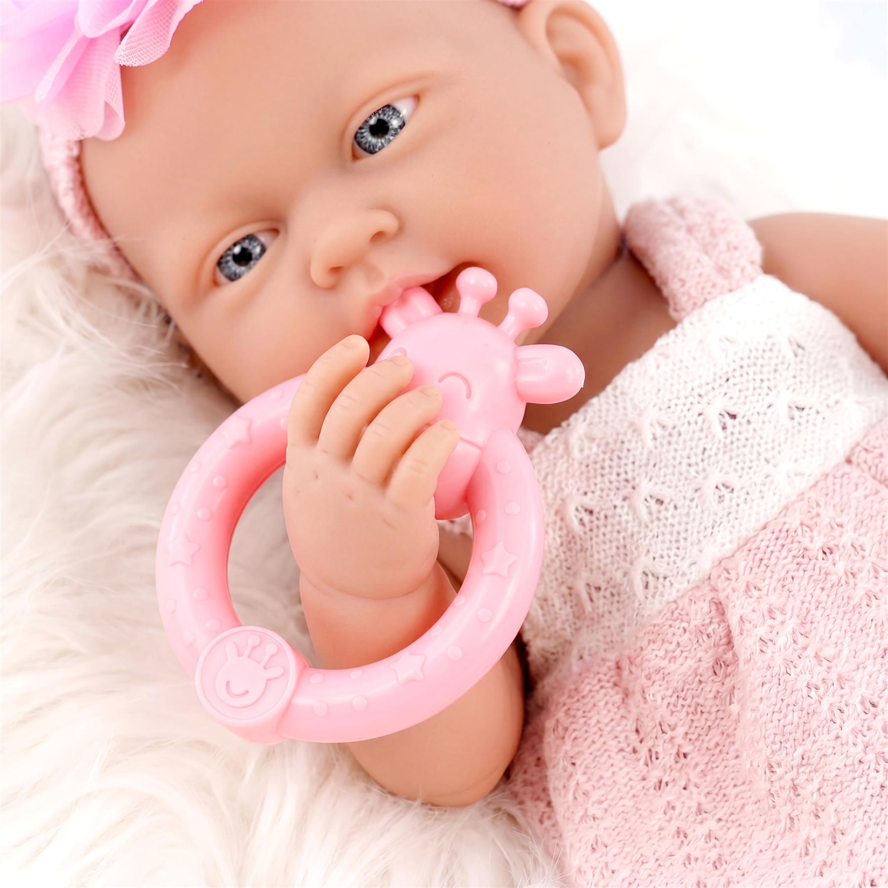 Blue-eyed baby doll in a pink outfit holds a ring-style pink pacifier toy.