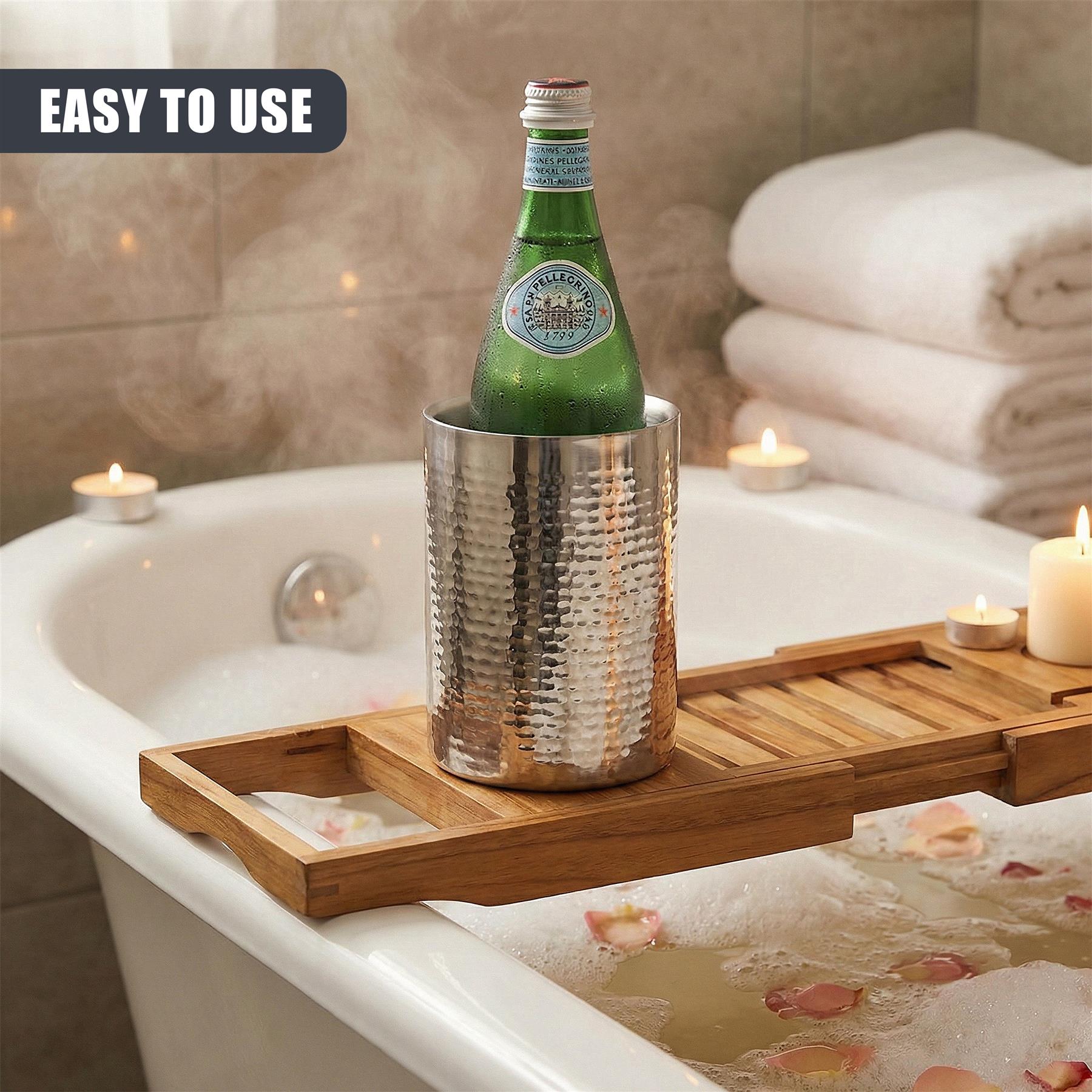Metal wine cooler and green bottle resting on a wooden tray in a bathtub.