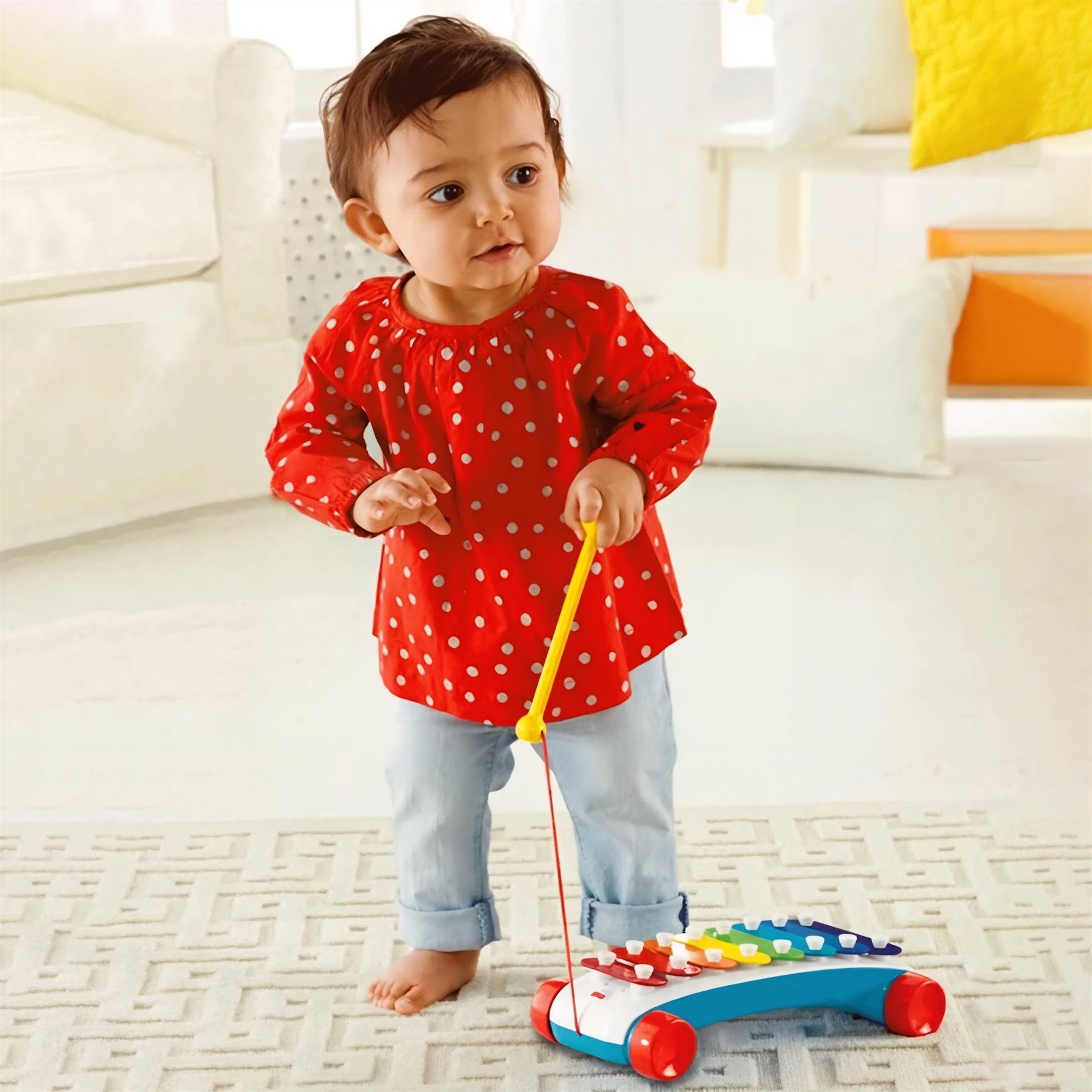 A toddler in red clothing plays with a colorful, xylophone-style musical pull toy on the floor.