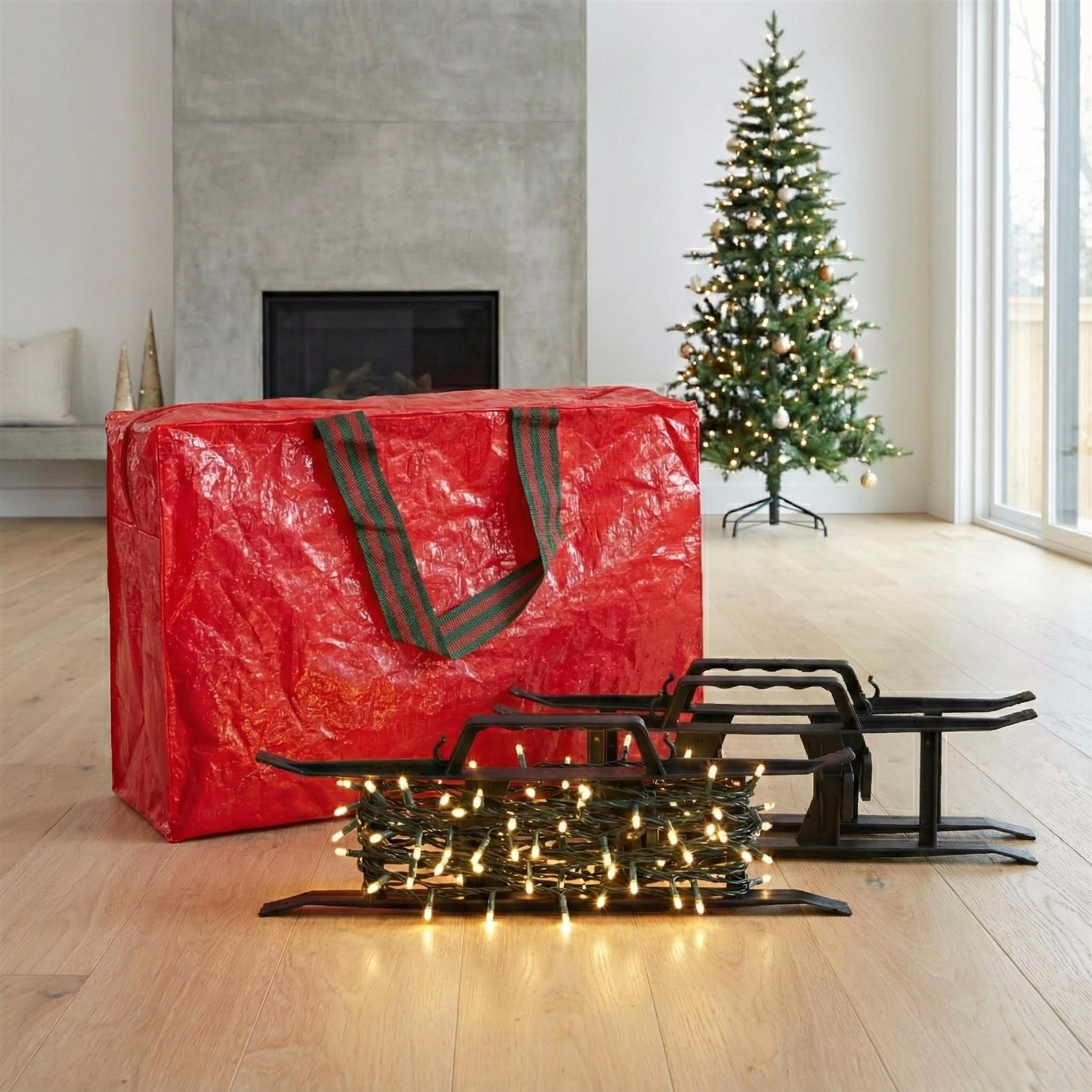 A red storage bag with a green and red striped handle sits on a wooden floor in front of a Christmas tree. The bag is filled with a set of Christmas lights and a set of Christmas decorations, including a string of lights, a set of Christmas lights, and a set of Christmas decorations. - The Magic Toy Shop