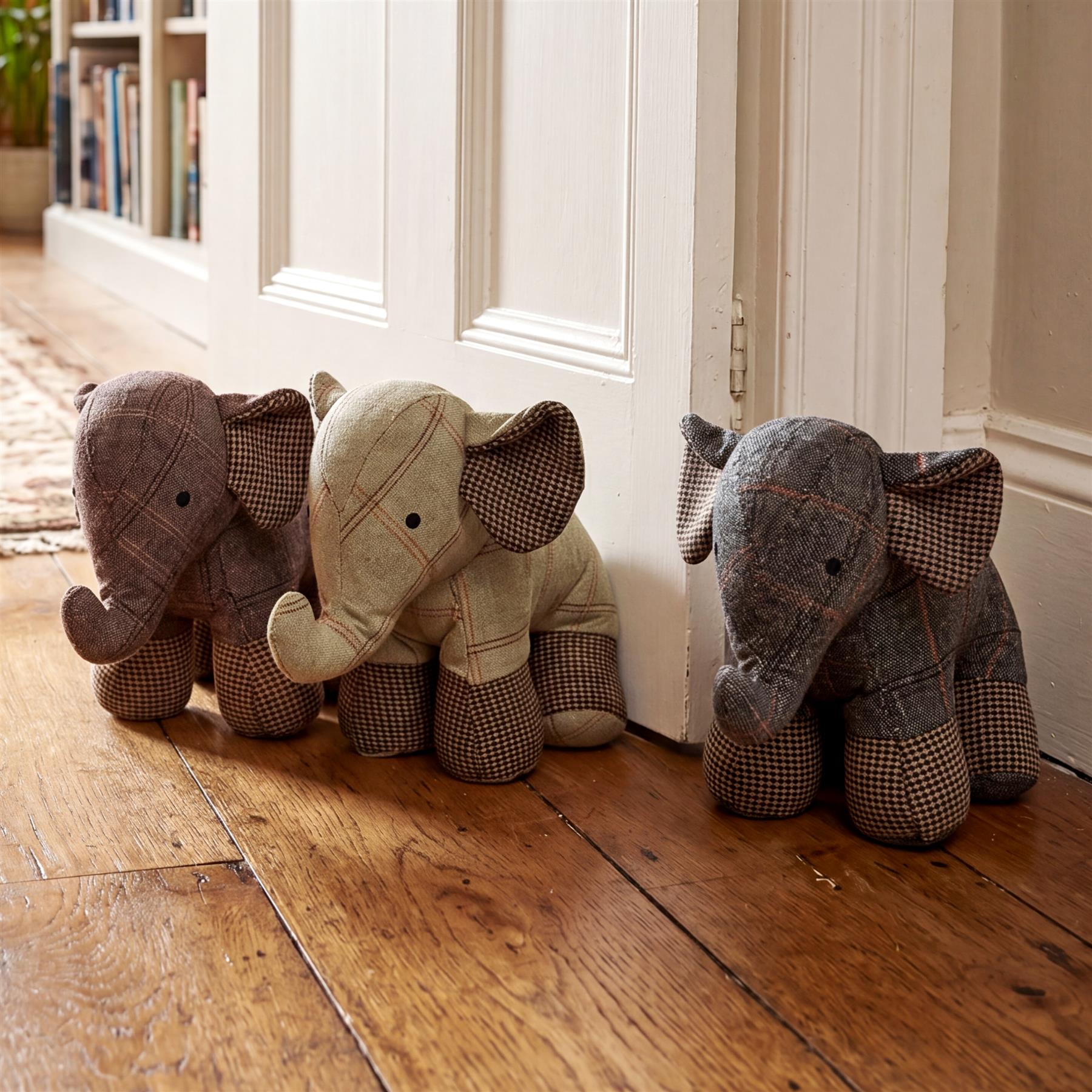 Three plaid stuffed elephants, used as door stoppers, rest on a polished wooden floor.