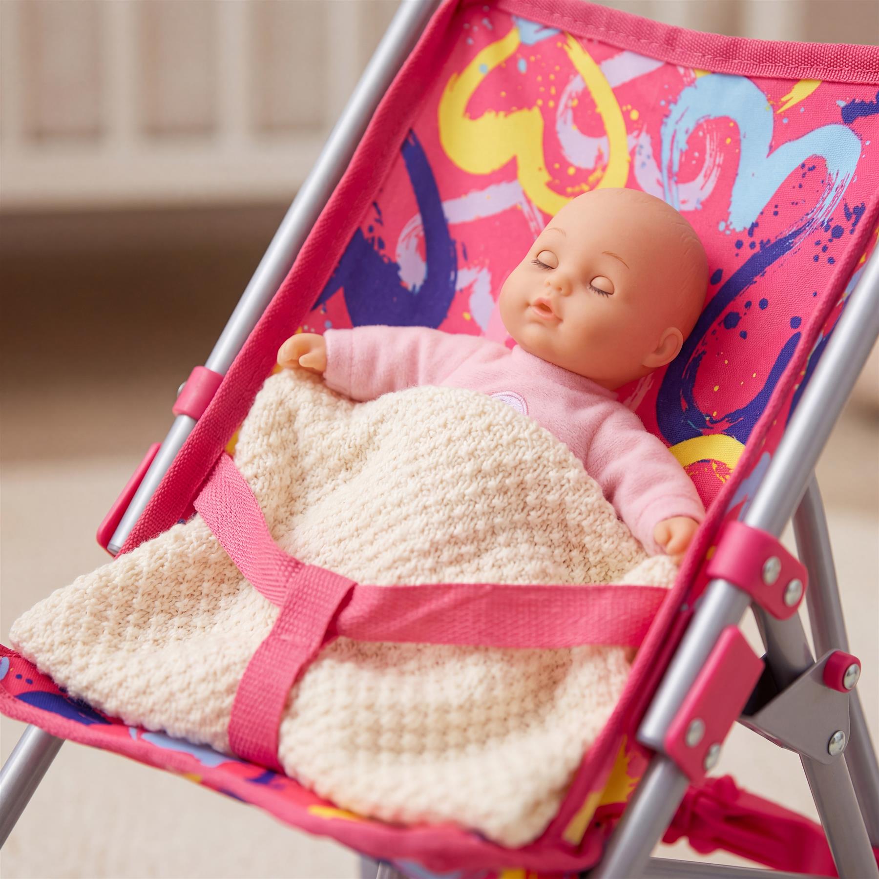 A doll wrapped in a beige knit blanket rests in a colorful pink patterned buggy stroller seat.