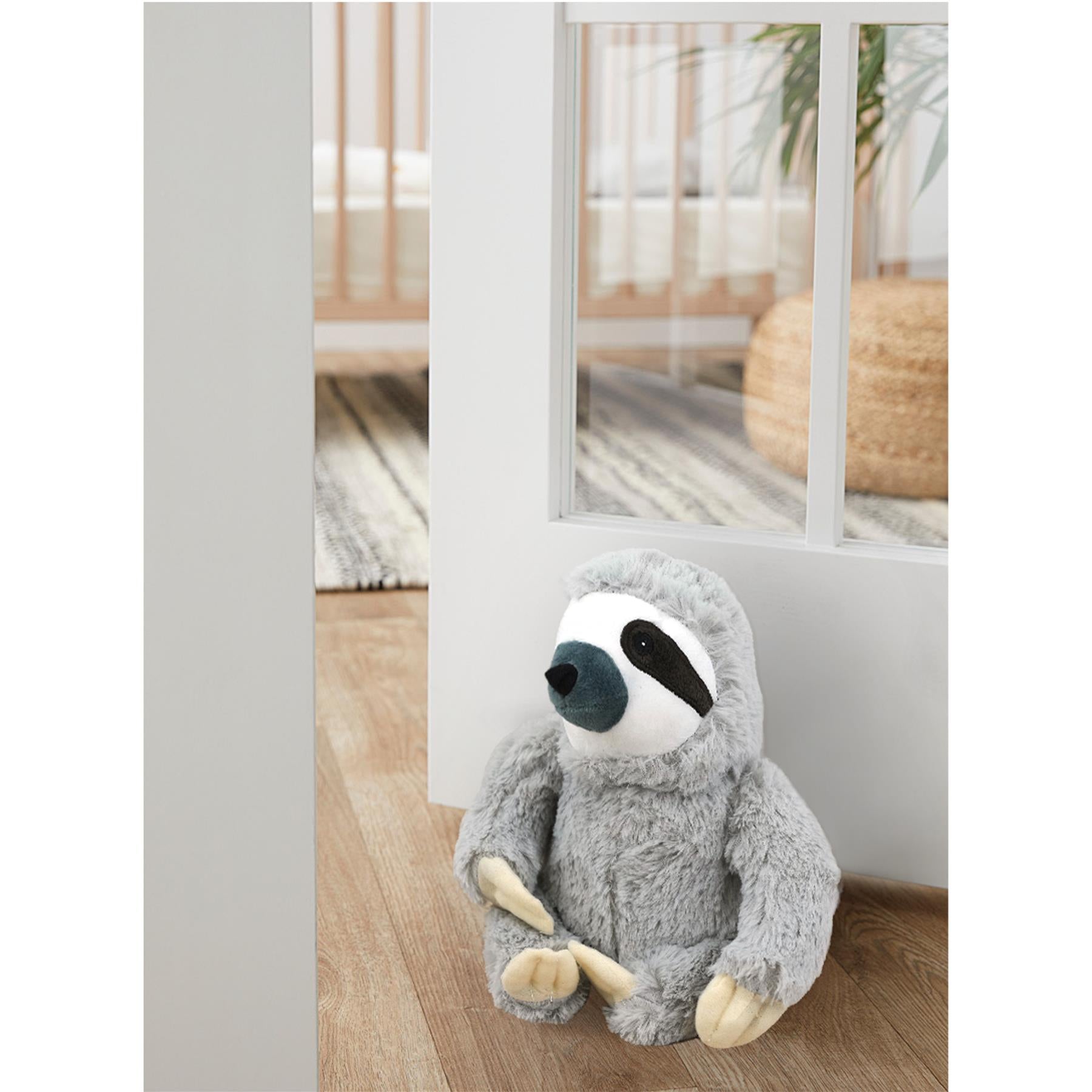 A plush sloth sitting on a floor, looking through a white door. The sloth has a soft grey fur, a white face, and black eyes. It has a few small toys around it, including a ball and a small plant. The background is a white wall with a few pieces of furniture, such as a chair and a table. The overall atmosphere is warm and inviting, with a sense of comfort and relaxation. - The Magic Toy Shop