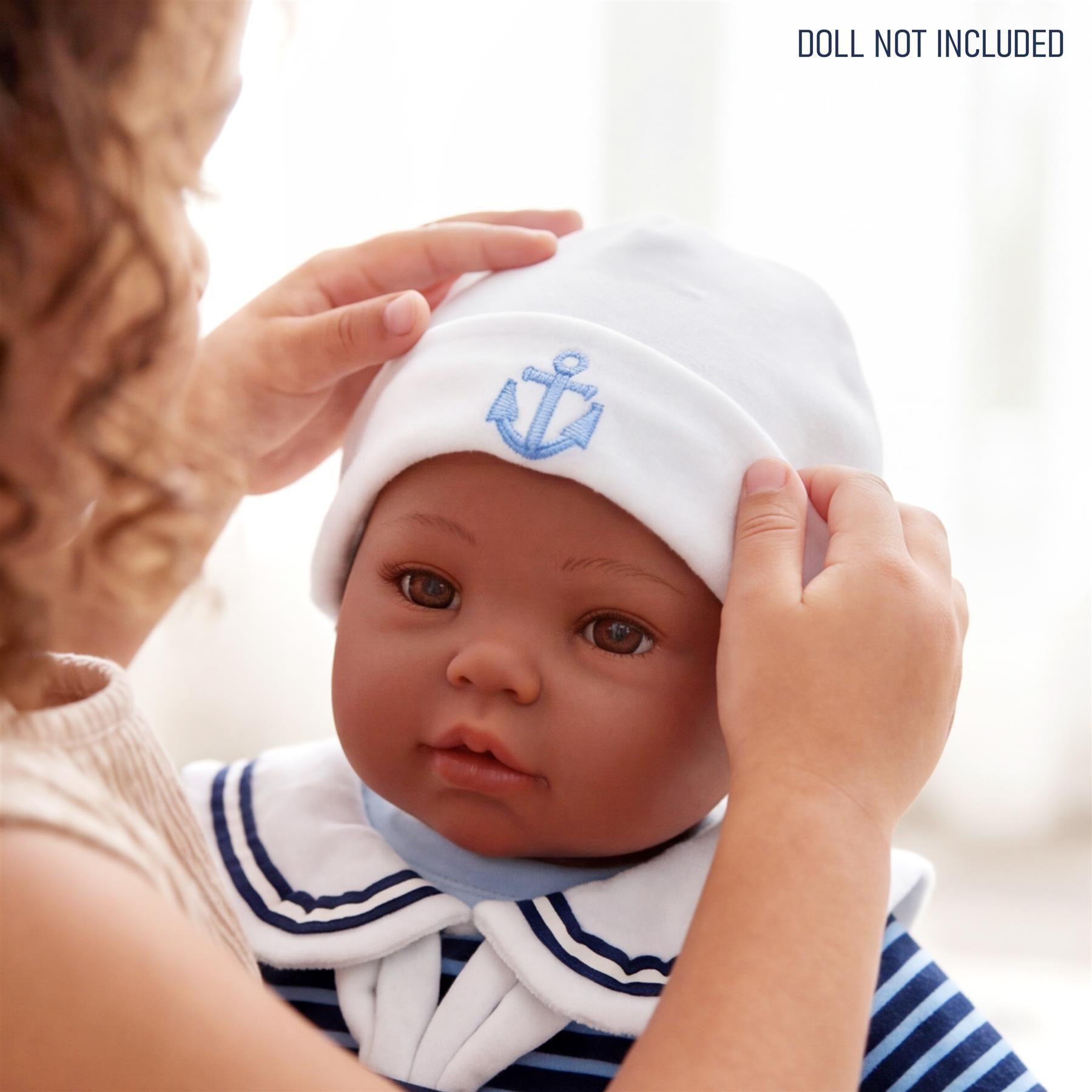 Hands place a white sailor hat featuring a blue anchor emblem on a reborn doll wearing a striped outfit.