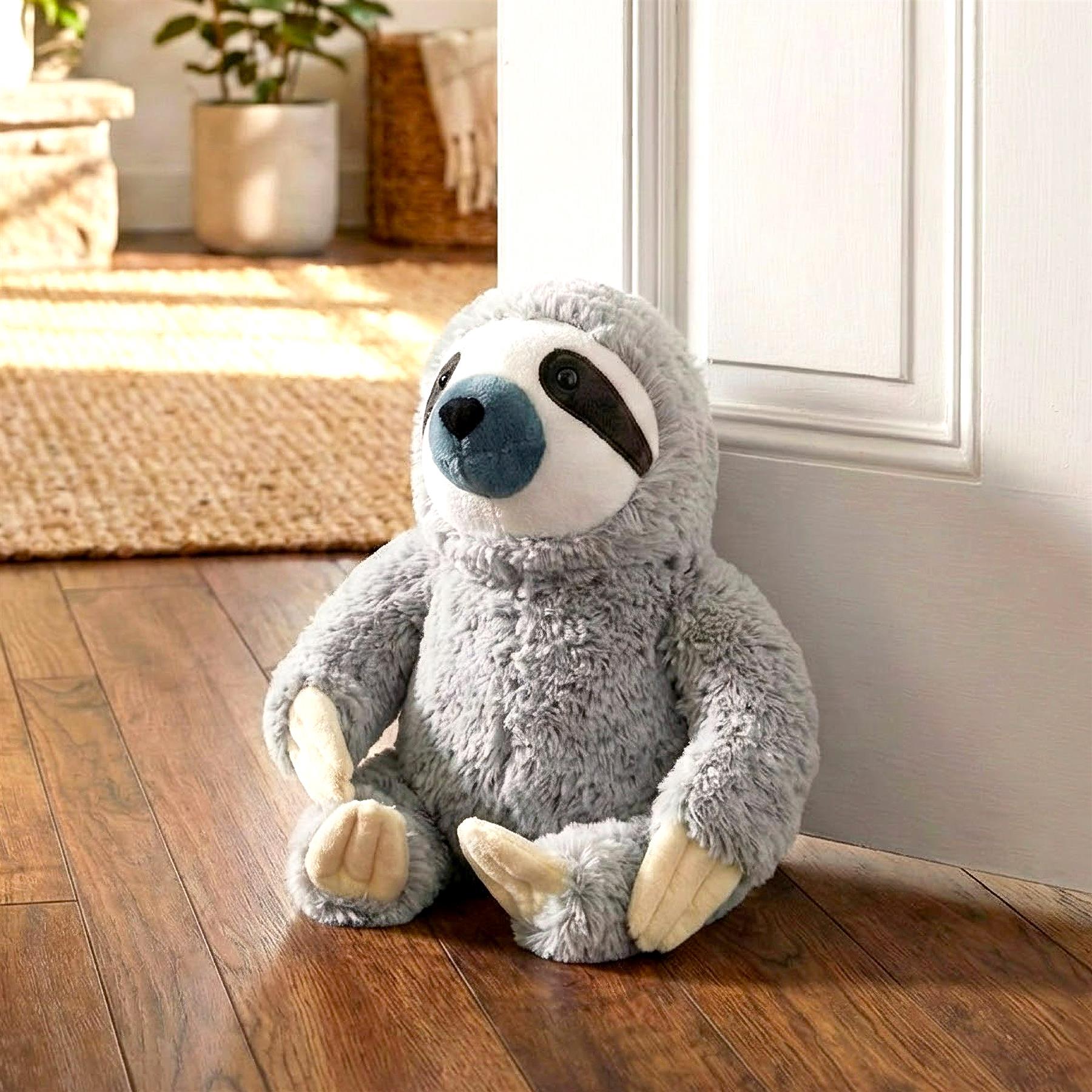 A cuddly sloth plushie sitting on a wooden floor, looking directly at the camera. The plushie is made of soft, grey fur with white and blue markings on its face. Its arms and legs are white and it has a blue nose. The background is a warm, sunlit room with a woven rug, a potted plant, and a wicker basket. - The Magic Toy Shop
