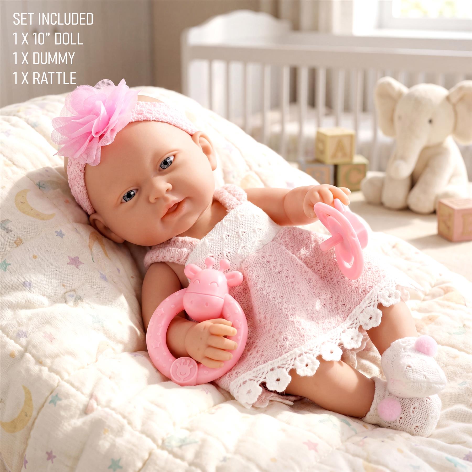 The doll lies on a patterned blanket with a pink rattle and flower headband.