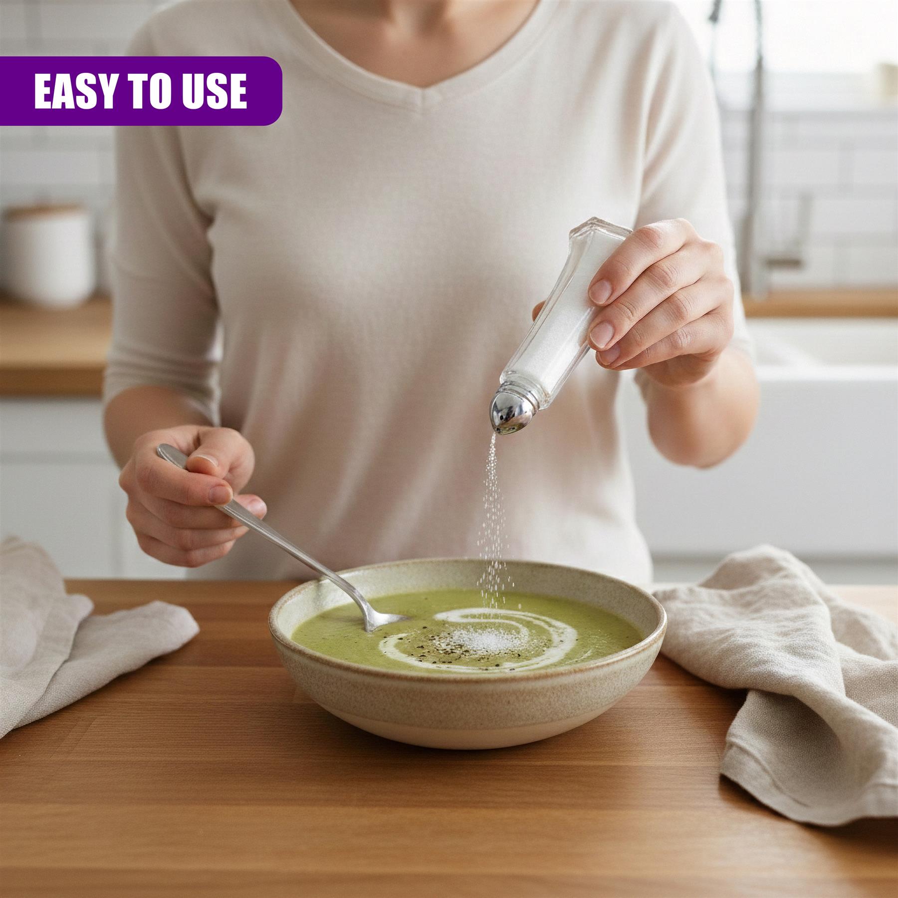 Hands sprinkle white seasoning from a glass shaker bottle into green soup served in a shallow bowl.