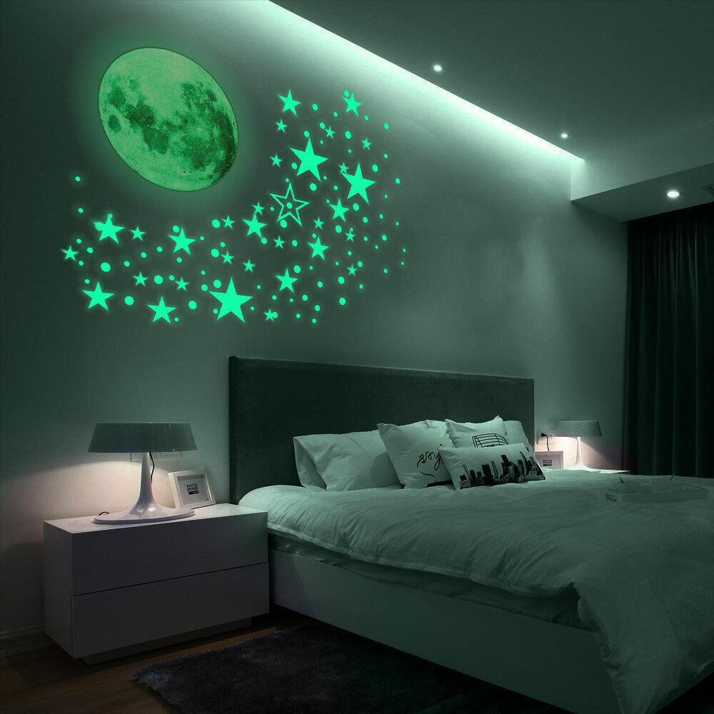 A bedroom at night with a glowing moon and stars on the wall. The bed is made with white sheets and pillows, and a dark grey headboard. A white nightstand with a lamp sits next to the bed. The room is dimly lit, with the only light coming from the glowing wall decorations. - The Magic Toy Shop