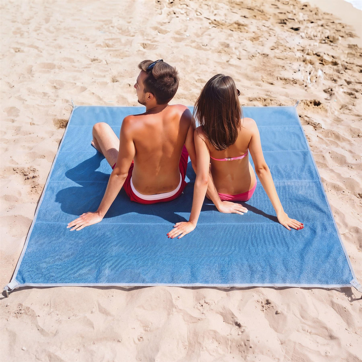 A couple sits on a blue GEEZY beach mat on a sandy beach