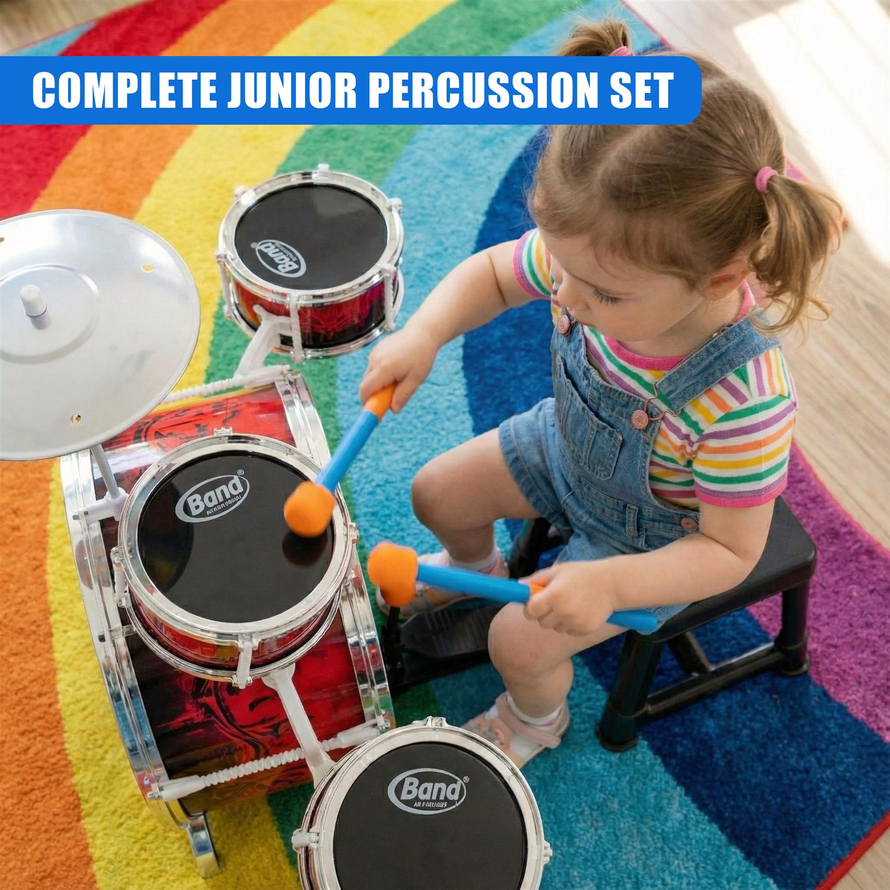 A young child plays a mini drum set on a colorful rug. The drum set has a red and black drum set with silver hardware and a white drum stool. The child is wearing a rainbow-striped shirt and denim overalls, and is holding two drumsticks. The drum set is sitting on a bright, multi-colored rug with a rainbow pattern. The child is sitting on a black drum stool, playing the drums. - The Magic Toy Shop