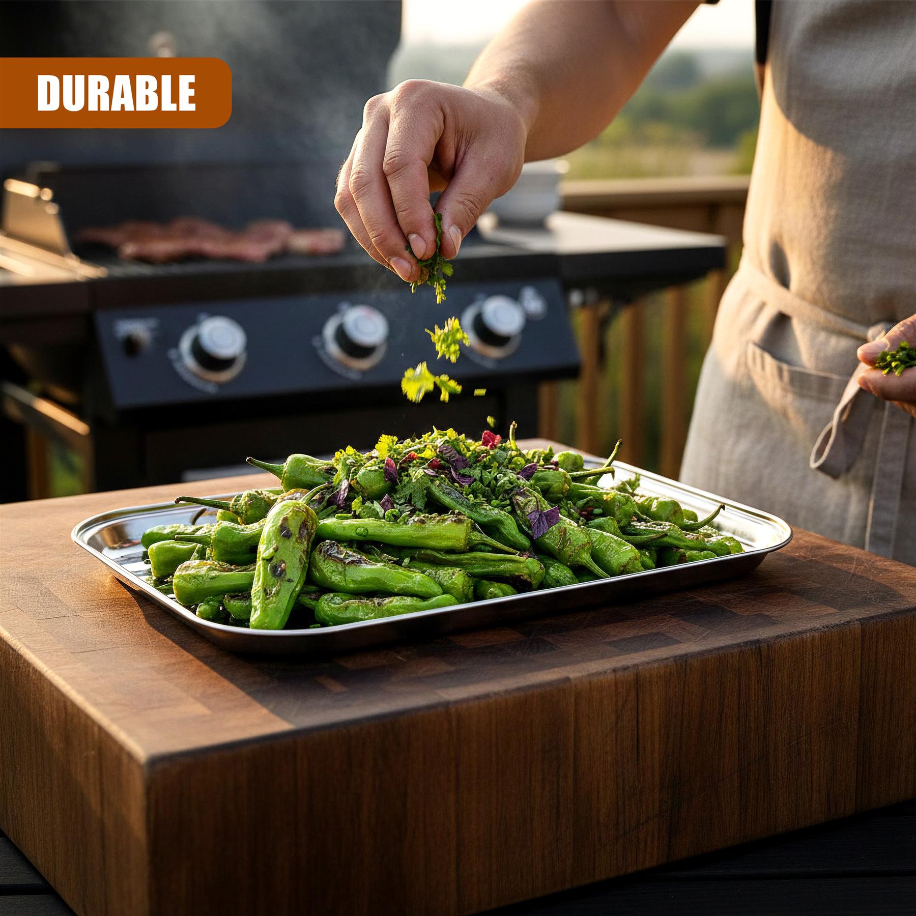Hand sprinkles chopped green herbs over grilled vegetables arranged on a rectangular metal tray.
