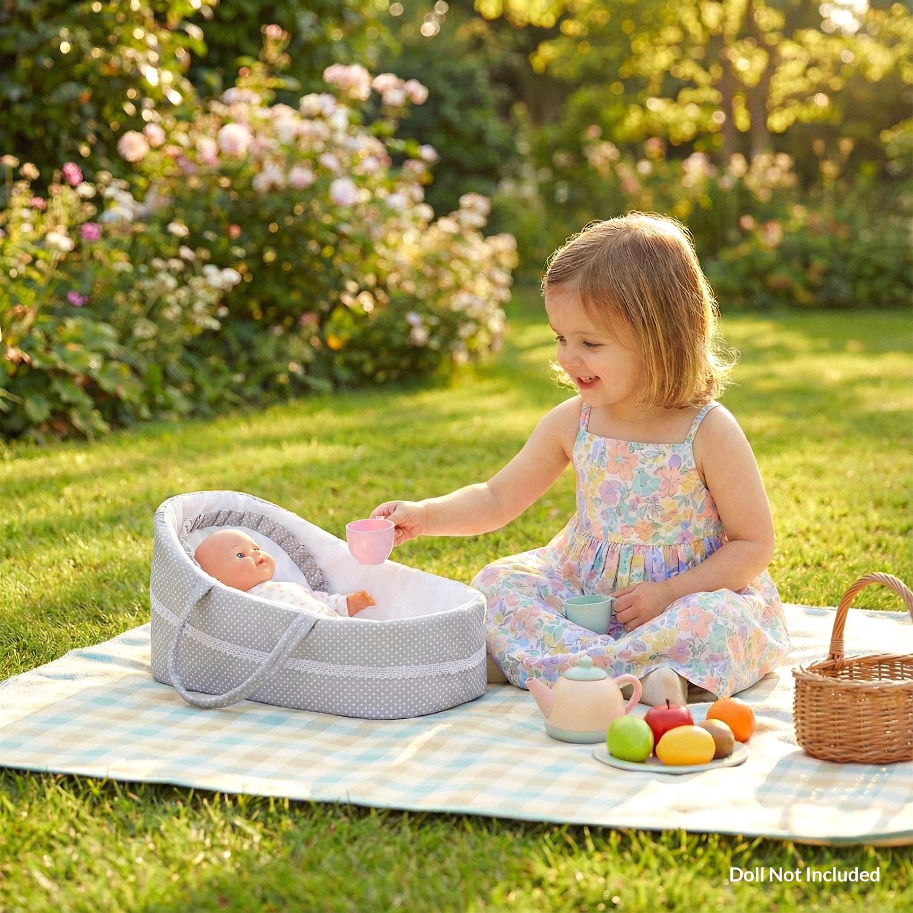 A child plays with a doll and a baby doll in a garden. The child is sitting on a picnic blanket, with a doll in a bassinet, a teapot, and a basket of fruit. The child is holding a cup and smiling at the doll. The scene is set in a garden with flowers and trees in the background. - The Magic Toy Shop