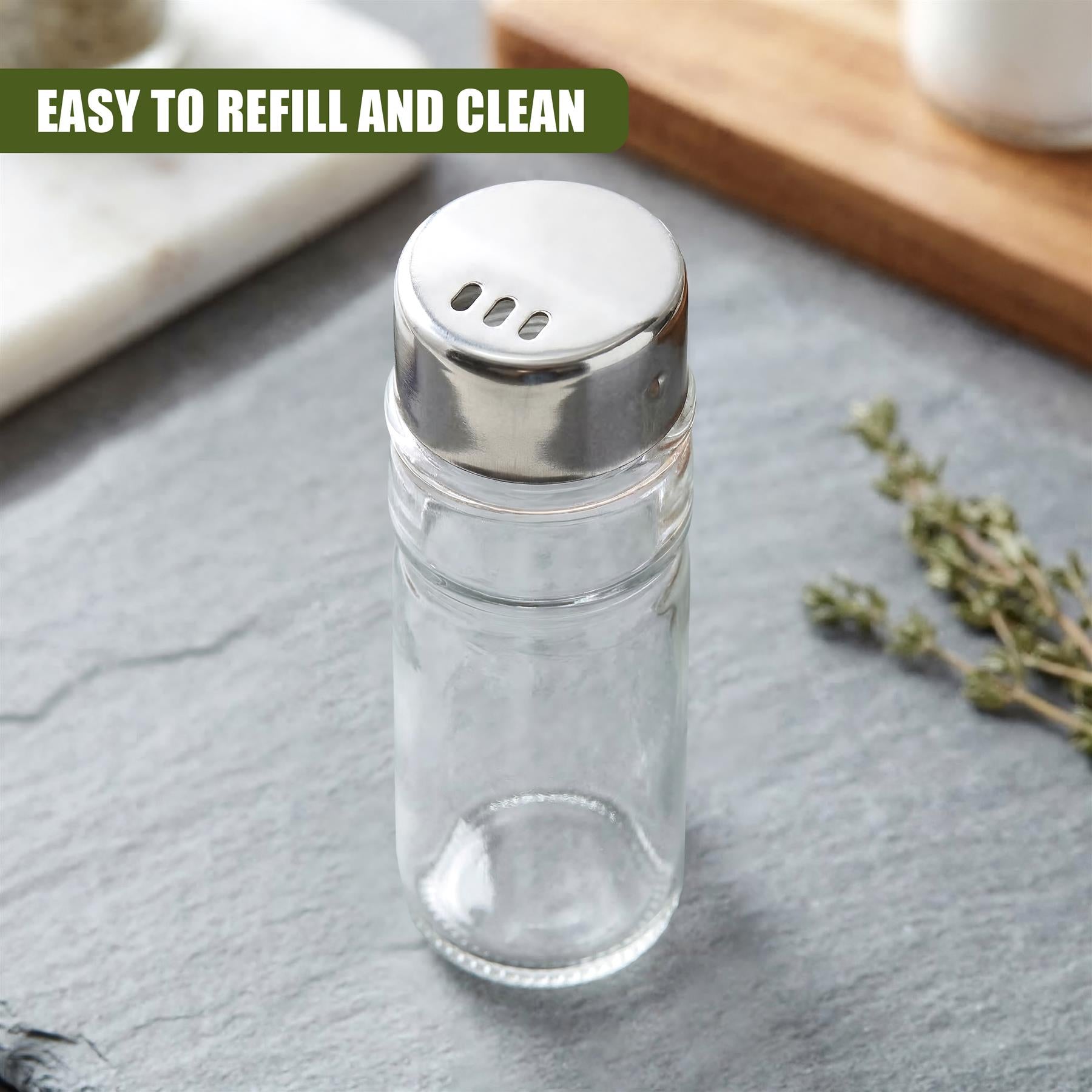 A clear glass spice jar with a silver lid sits on gray slate next to sprigs of dried herbs.