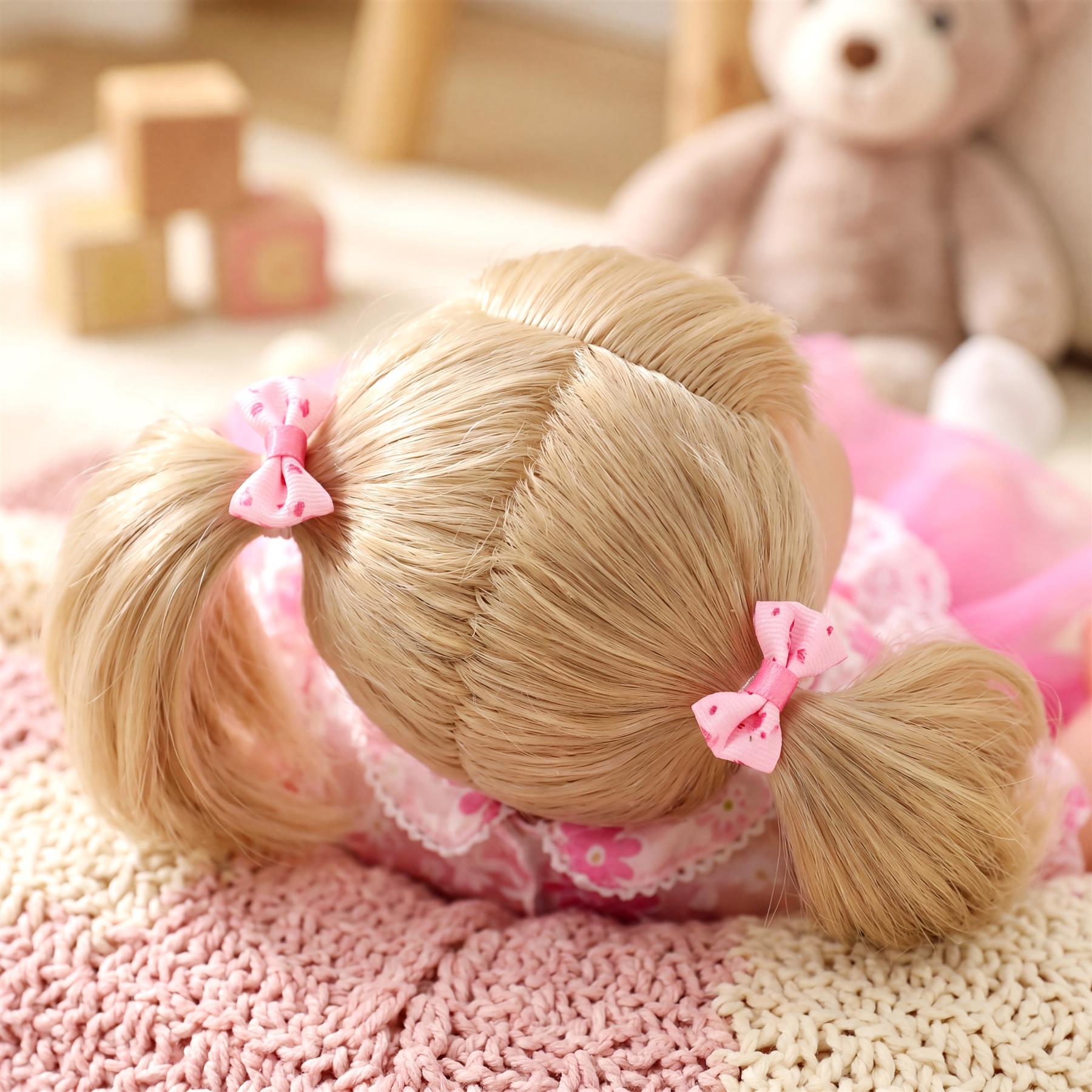 Blonde baby doll with braided pigtails and pink dress lies on soft pink blanket.
