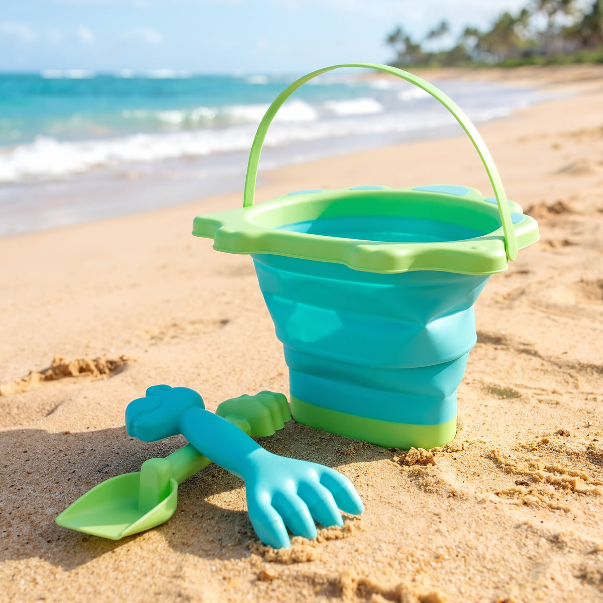A teal and green collapsible bucket and spade set sits on a sandy beach
