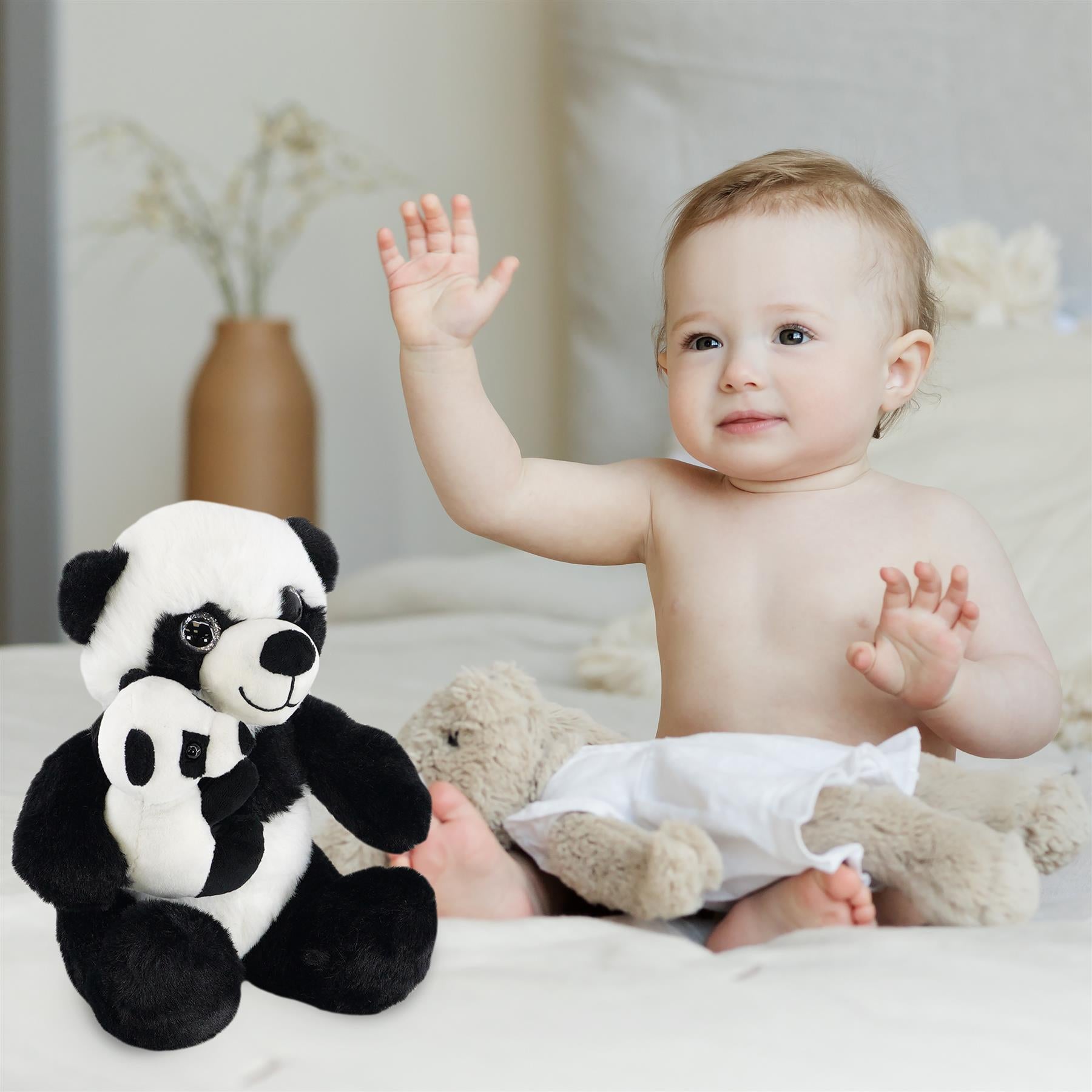 A joyful baby sits on a soft bed, raising one hand while smiling at the camera. Next to the baby is a super soft mommy panda plush toy holding a smaller panda, with a beige teddy bear companion nearby. The warm, cozy setting highlights the cuteness of both the baby and the plush toys. Perfect for cuddles and playtime, this adorable set is ideal for young children. themagictoyshop