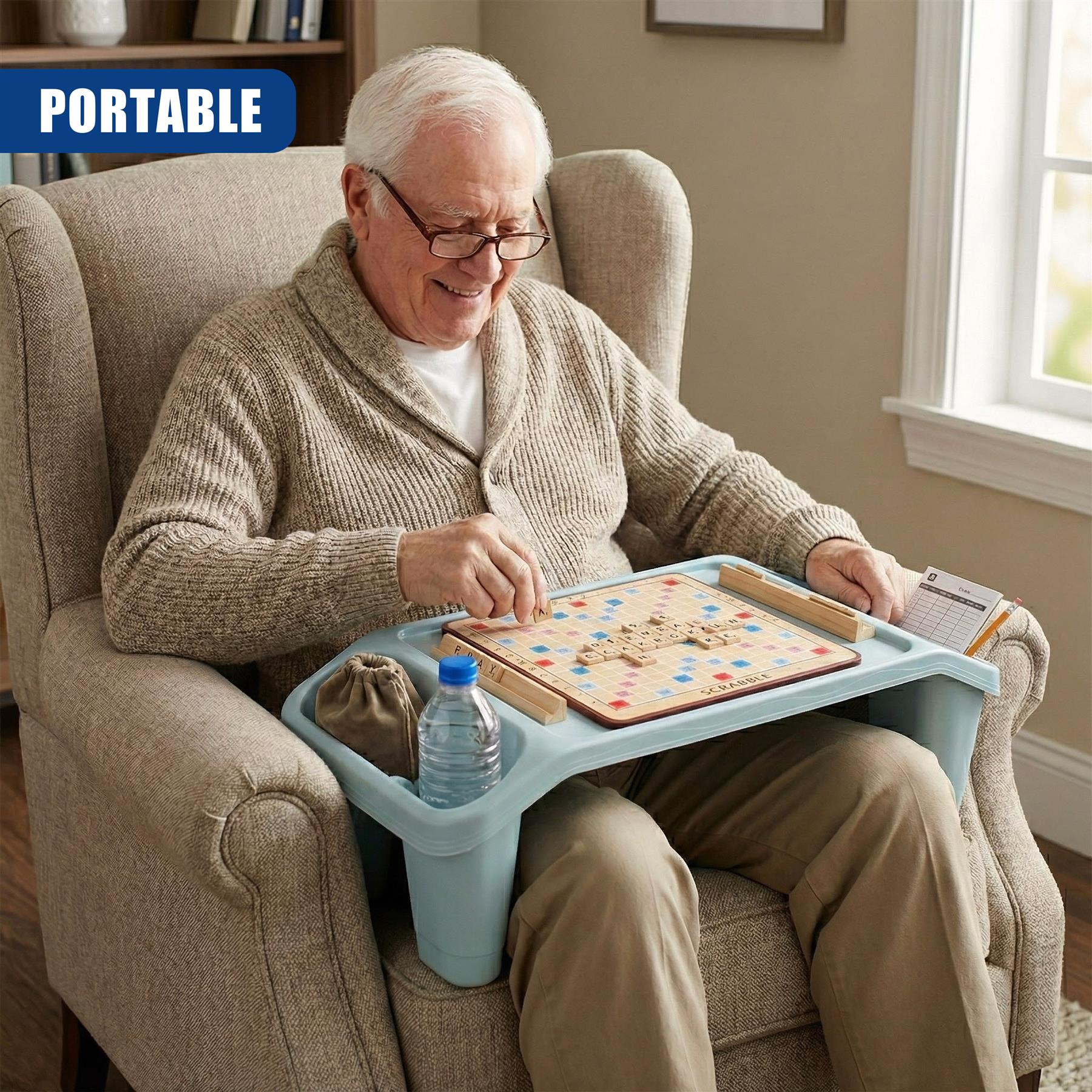 A senior man with white hair and glasses, sitting in a beige armchair, playing a Scrabble game on a portable, foldable table. The table is blue, with a built-in cup holder and storage compartment, and has a Scrabble board, tiles, and a water bottle on it. The man is smiling, and the room is sunny with a window in the background. - The Magic Toy Shop