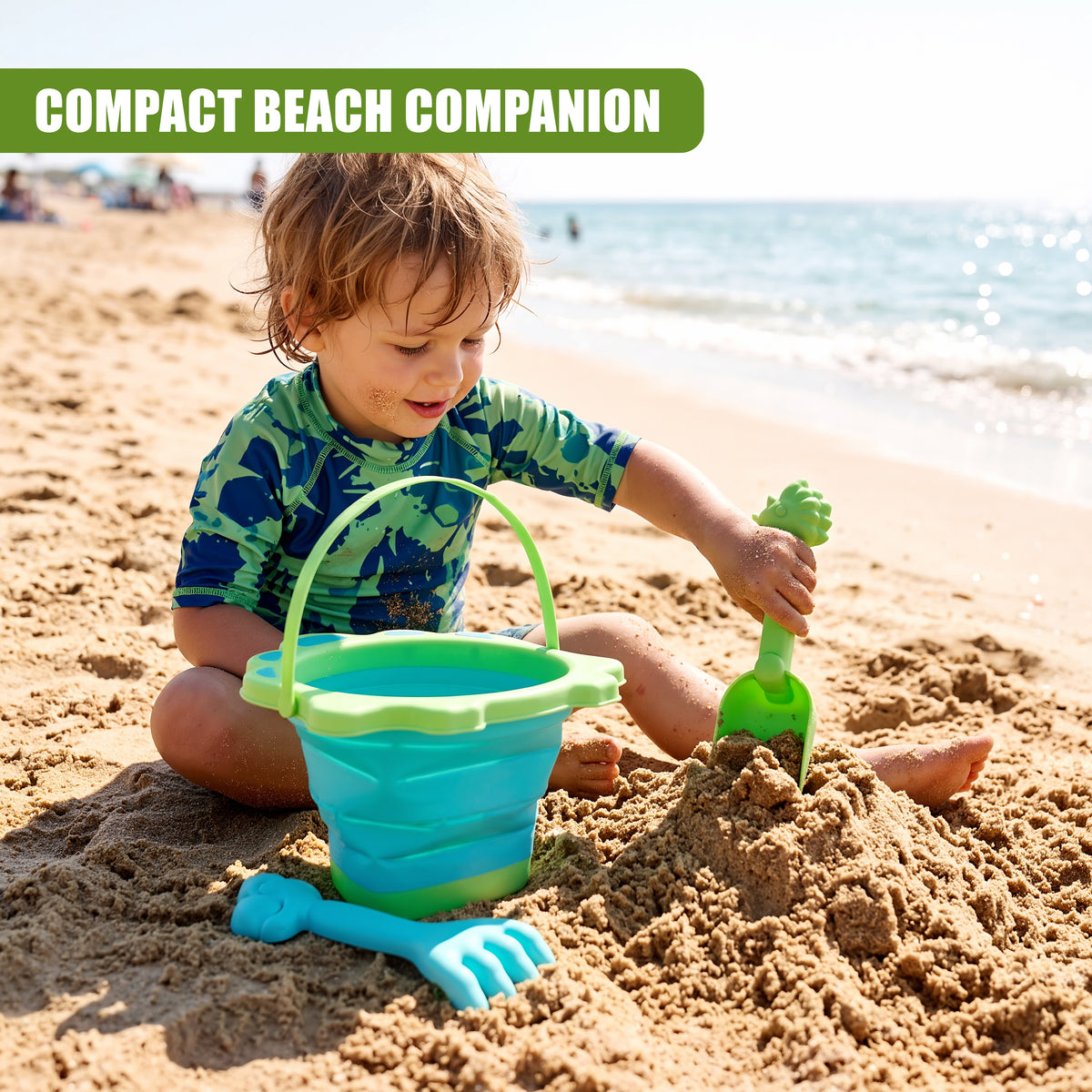 A young boy plays with a blue and green collapsible beach bucket and spade on a sandy beach