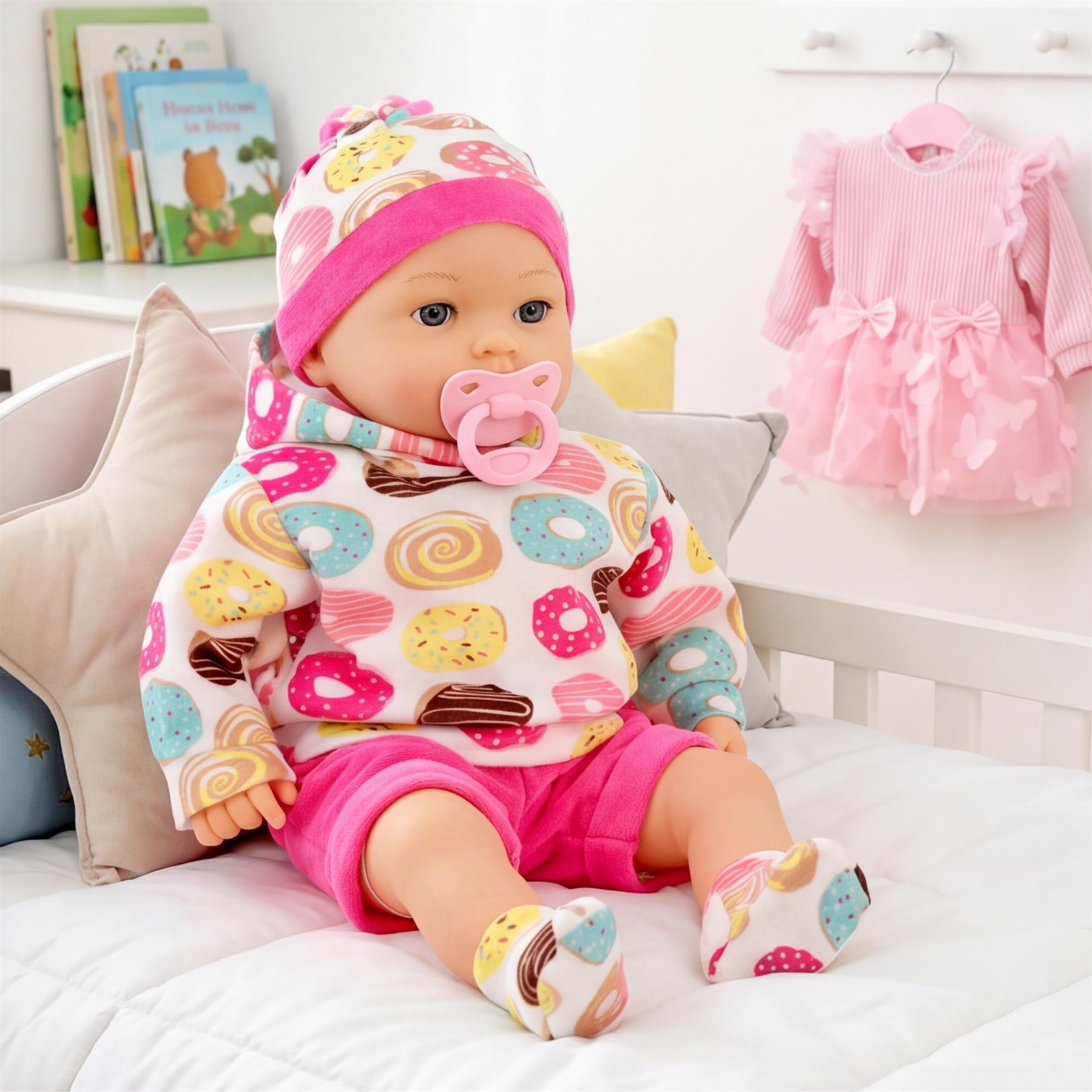 Doll Maeve sits on a bed wearing a patterned pink and white sleeper outfit, hat, and pacifier.