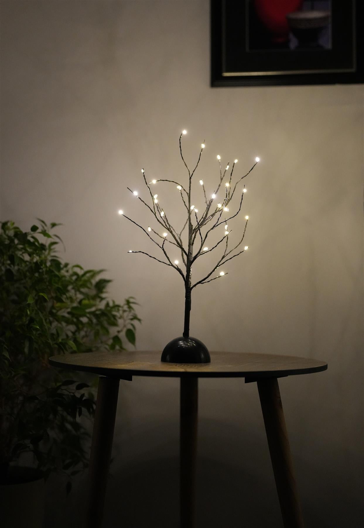 A decorative LED tree lamp with 32 warm white lights, designed with slender branches and a round black base, placed on a wooden table. The background features a softly lit room with a green plant and framed artwork. Ideal for home decor, ambient lighting, or as a unique gift. themagictoyshop.